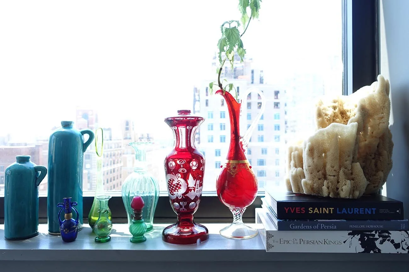 Decorative glass vases and bottles, a stack of books, and a large sponge-like sculpture on a windowsill with a city view in the background.