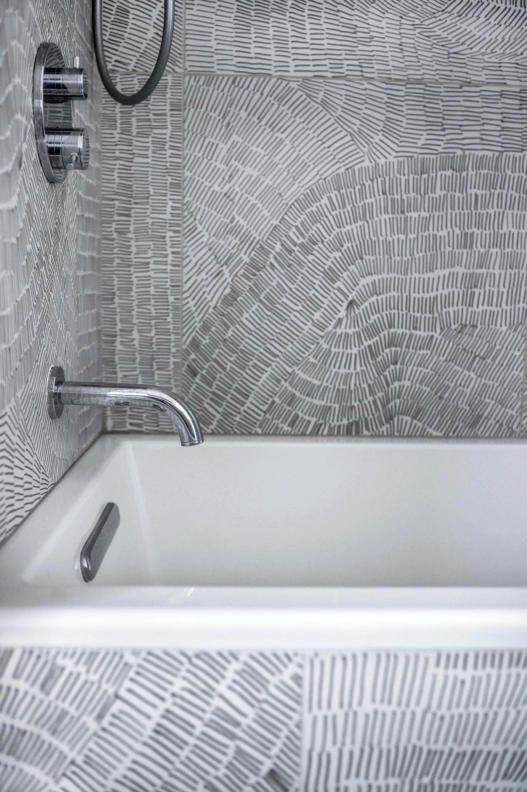 Close-up of a modern bathroom shower with black and white line patterned tiles, silver shower controls, and a white bathtub.