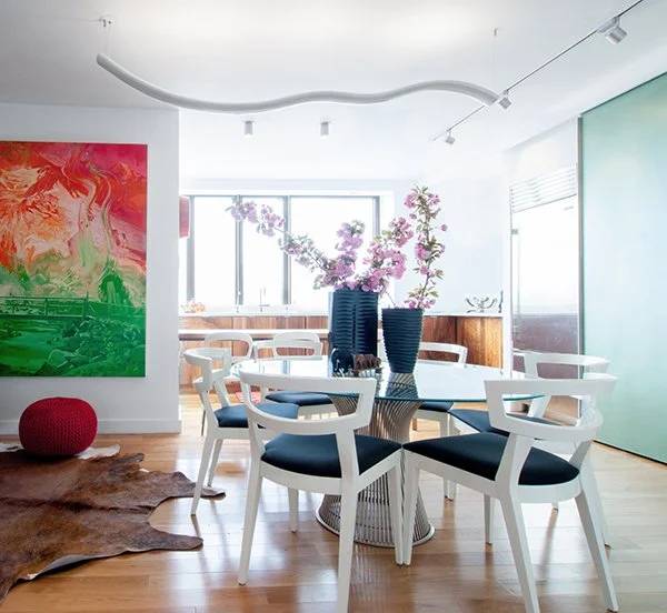 Modern dining room with a round table, six white chairs with black cushions, and a centerpiece of pink cherry blossoms in black vases. Large window, abstract painting, cowhide rug, and wood floor.
