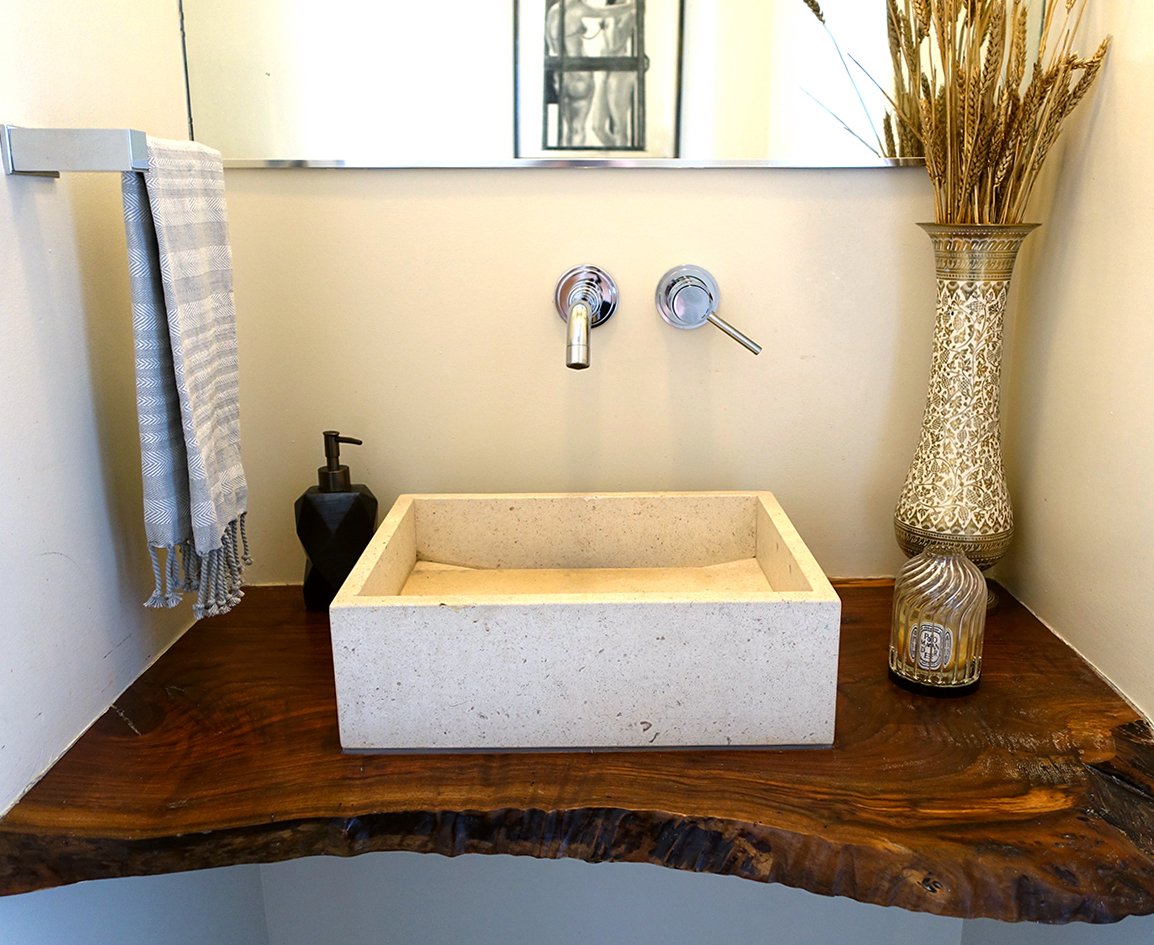 Bathroom vanity with a rectangular stone sink, a soap dispenser, a towel on a rack, a large decorative vase with dried flowers, a mirror above, and a reflective wall.