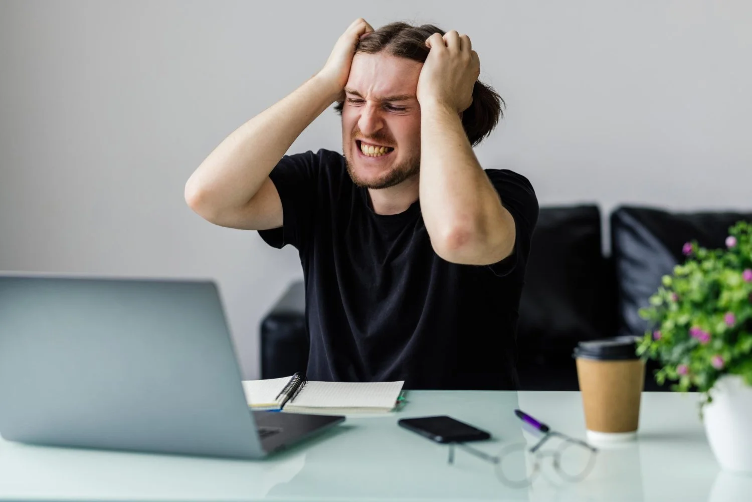 A young man with brown hair and a beard, wearing a black t-shirt, sitting at a white desk with a gray laptop, a notebook, a smartphone, glasses, a pen, a disposable coffee cup, and a potted plant. He appears stressed or frustrated, holding his head with both hands.