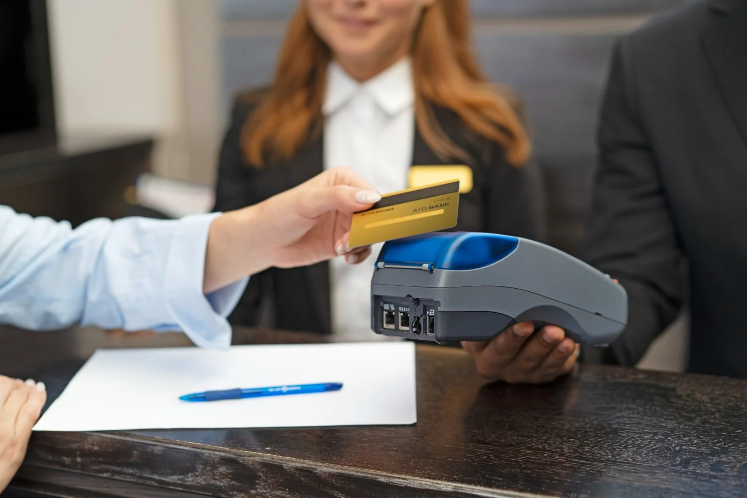Person swiping a yellow bank card through a card reader while standing at a desk, with a woman in a black blazer and white shirt in the background.