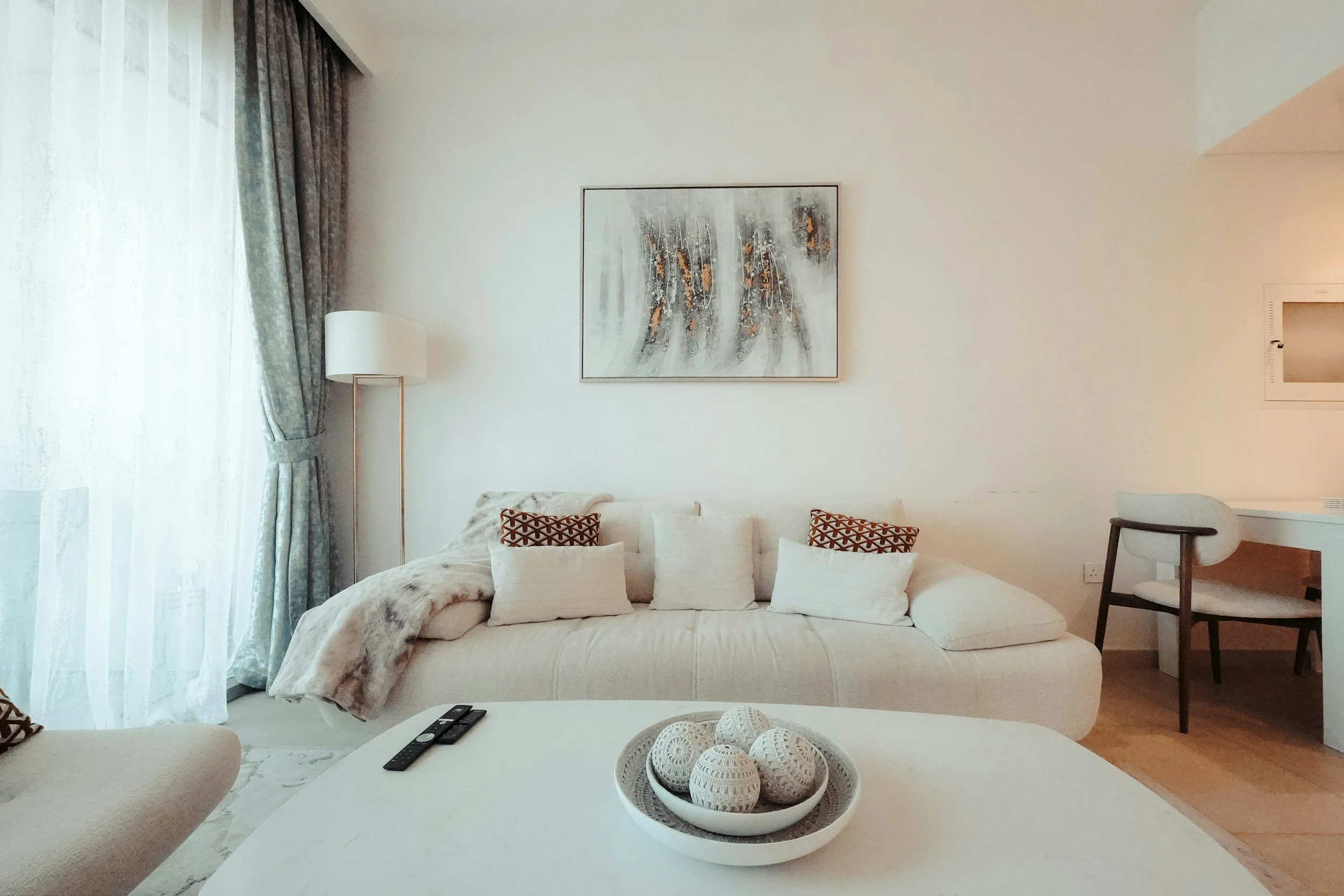 Living room with a beige sofa, decorative pillows, a white coffee table with a bowl of decorative balls, a large window with sheer curtains, a floor lamp, and a wall-mounted painting.