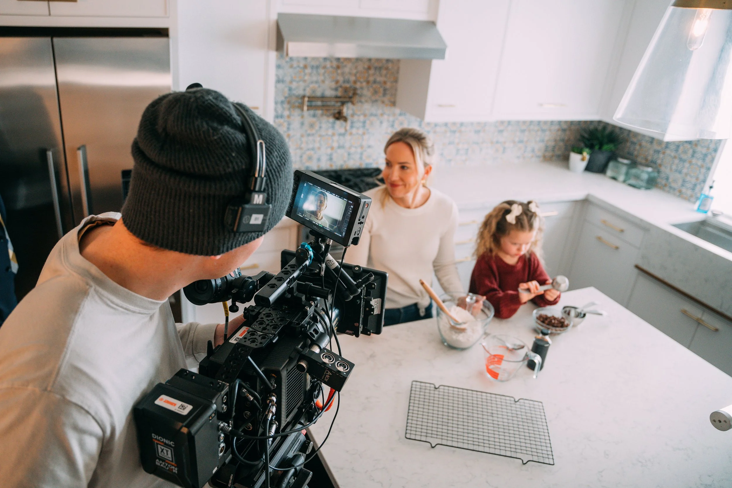 A woman and a young girl in a kitchen preparing food, filmed by a person with a camera and headphones.