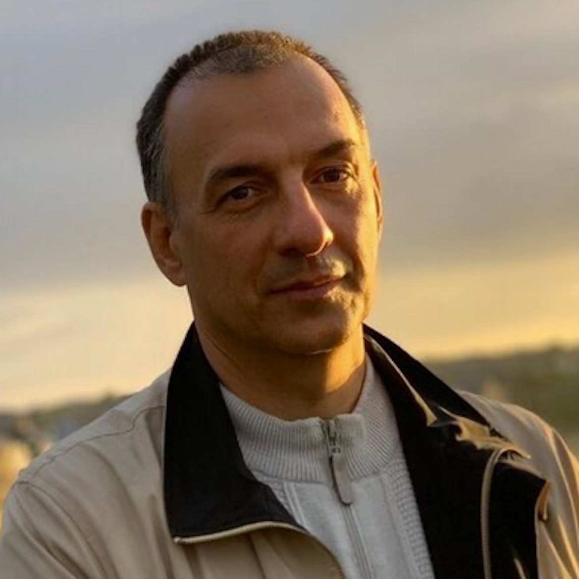 Uomo con giacca beige e sfondo di cielo al tramonto.