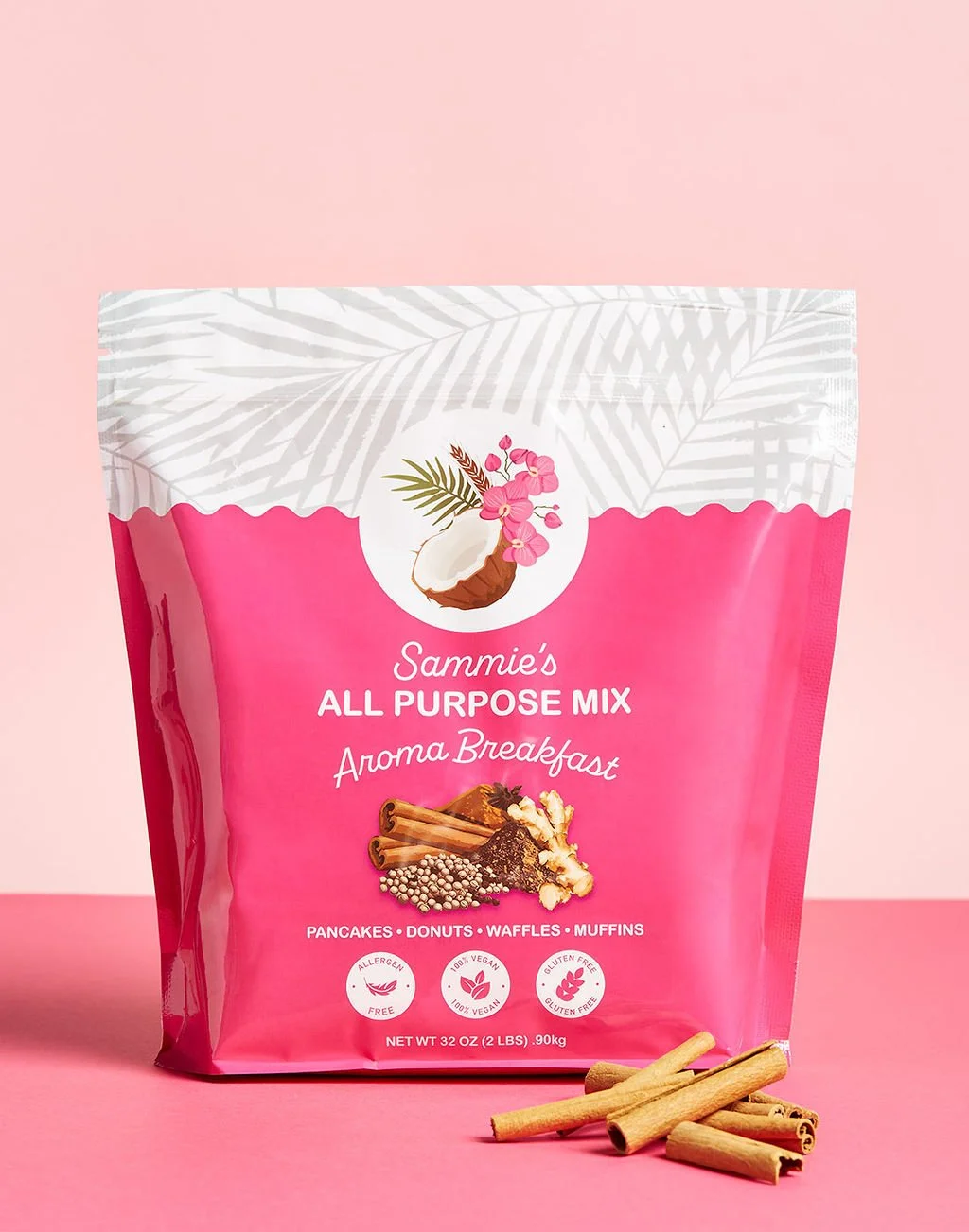 Pink packaging of Sammie's All Purpose Mix with illustration of coconut, pink flowers, and spices, placed on pink surface with cinnamon sticks in front.