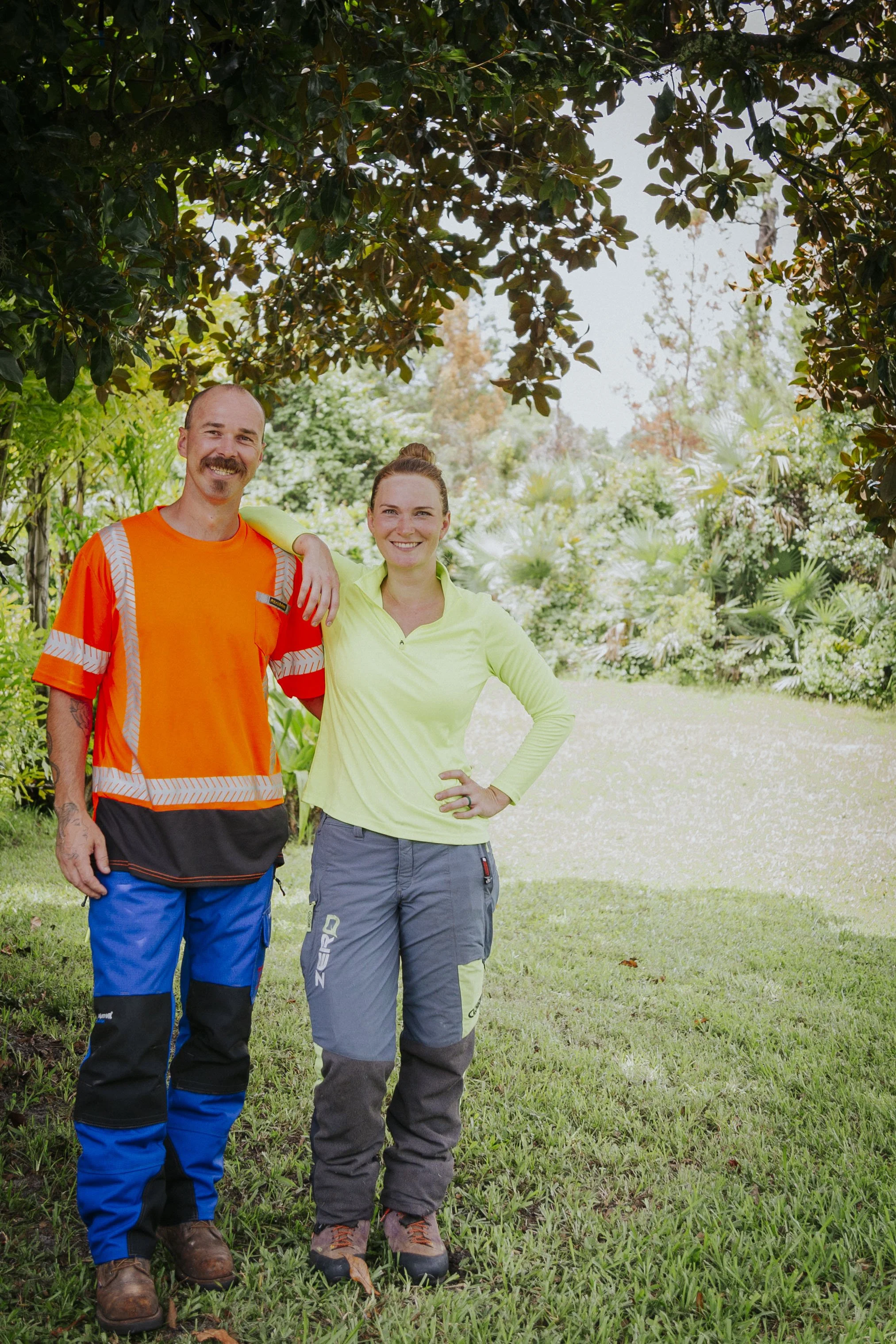 Two people standing outdoors on grass, smiling, surrounded by lush green plants and trees, with a garden background.