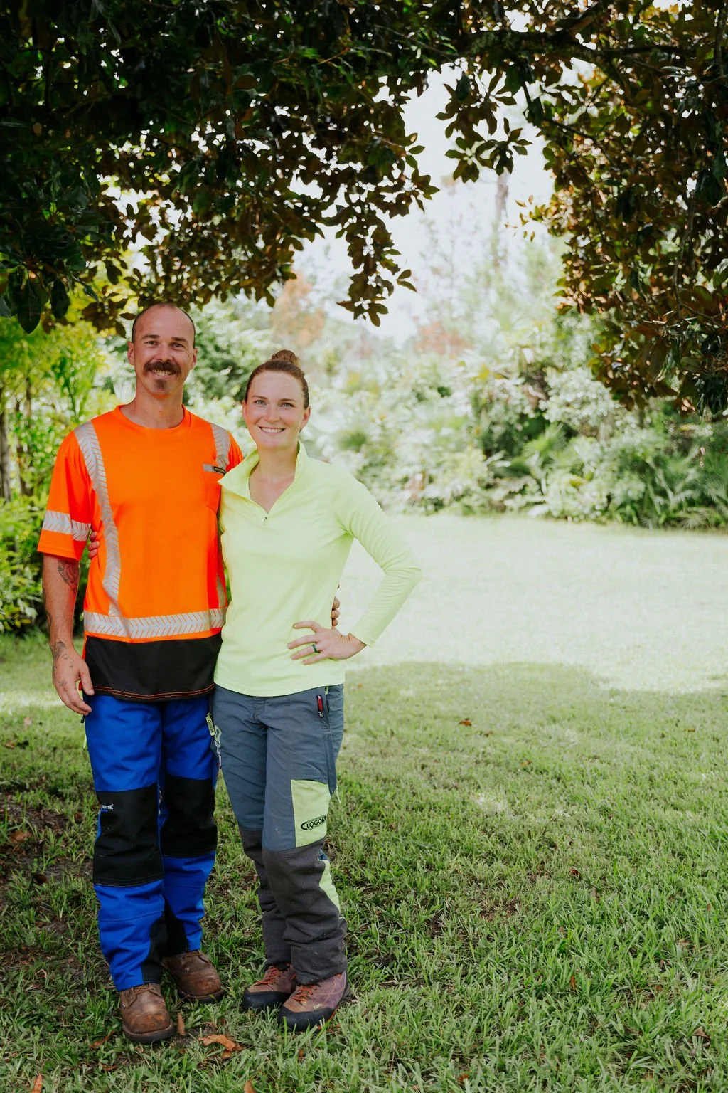 A smiling man and woman standing outdoors under a large tree in a park or garden, dressed in casual and work clothing, with lush greenery and plants in the background.
