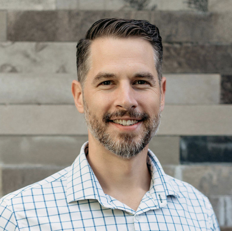 Noah Riner, Advisor at Hudson Street Ventures. A man with short dark hair, a beard, and a mustache smiling at the camera, wearing a white button-up shirt with a blue grid pattern. 