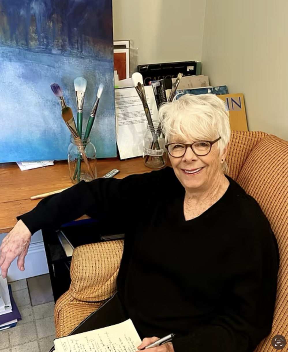 A woman with short white hair and glasses is sitting in an orange patterned armchair. She is smiling and holding a pen and paper. Behind her is a desk with art supplies, including brushes in glass jars and a painting.