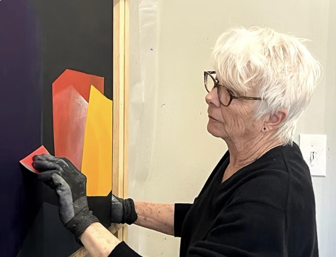 A woman with short white hair and glasses is hanging a piece of abstract art on a wall, wearing a black long-sleeve top and black gloves.