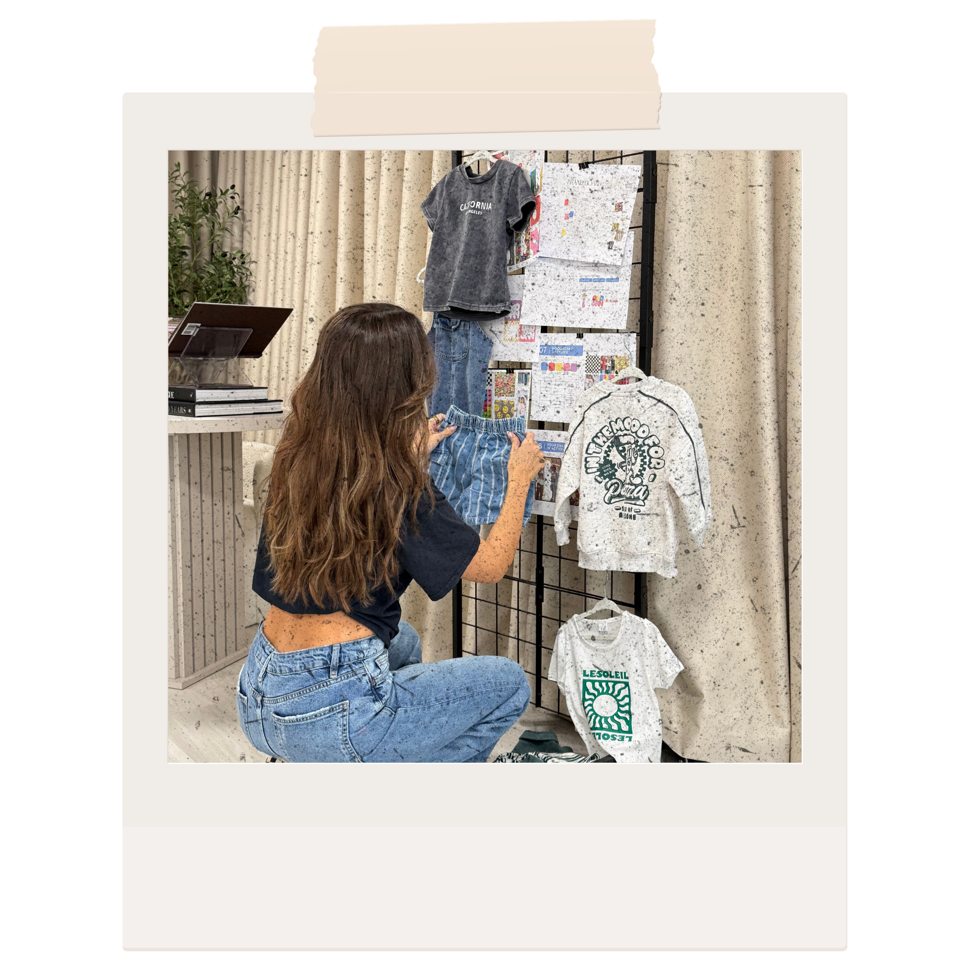 A person with long, wavy brown hair wearing a black T-shirt and ripped jeans is looking at merchandise, including t-shirts with logos and graphics, displayed on a black metal rack in a retail setting.