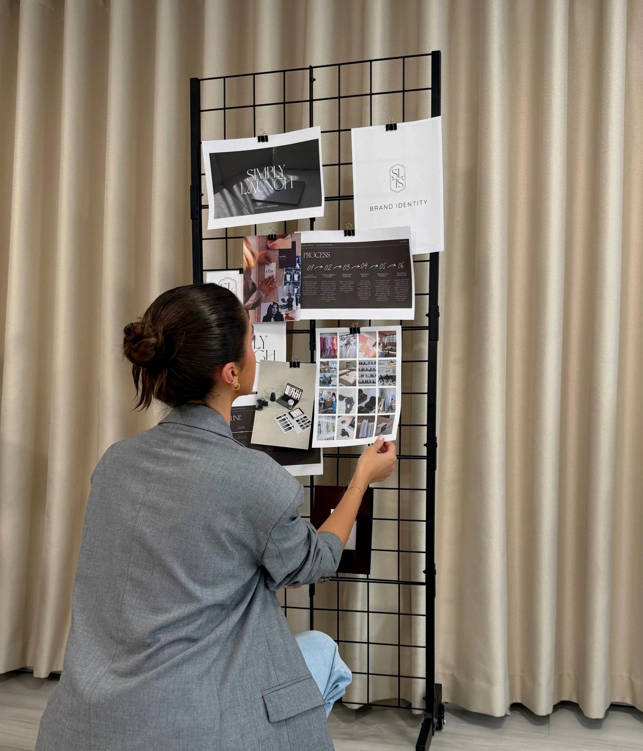 A woman in a gray blazer looks at a presentation board displaying branding and process diagrams at an indoor event, with beige curtains in the background.