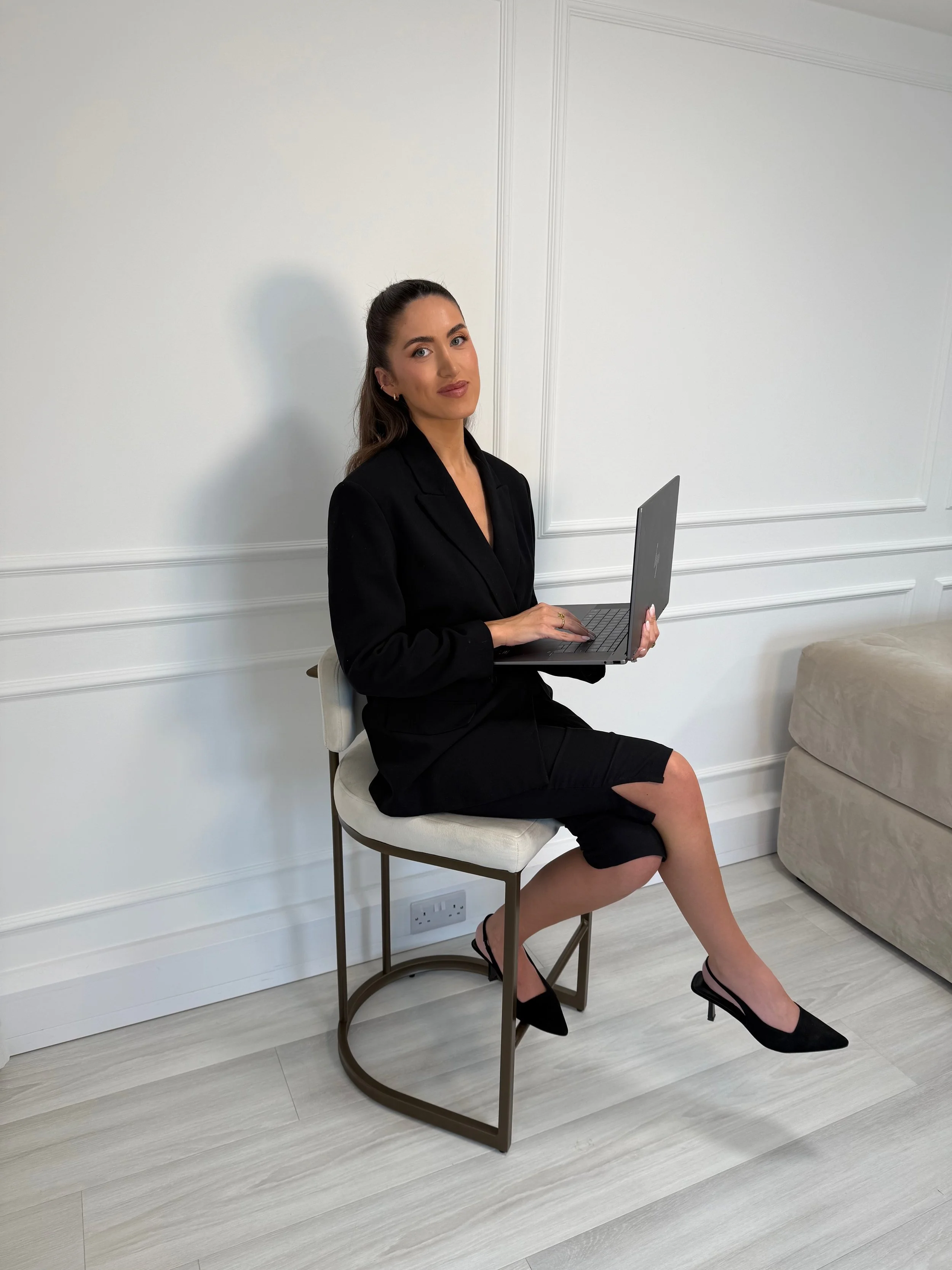 A woman dressed in a black blazer and skirt, sitting on a chair with a laptop in her lap, in a white room with wood flooring and a beige sofa to the right.