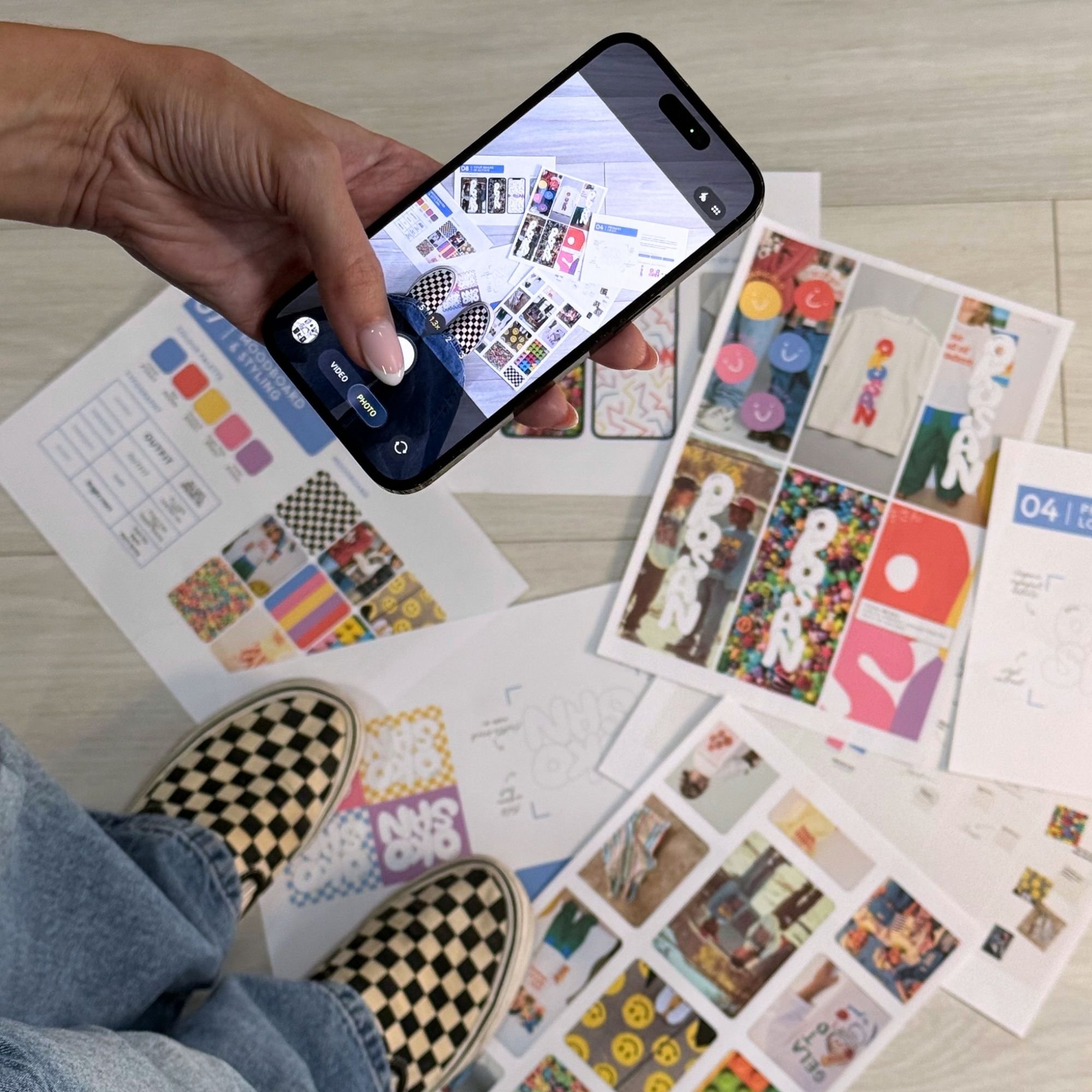 A person taking a photo of colorful sticker sheets with a smartphone, standing over flyers and papers on a table, wearing checkered slip-on shoes and jeans.