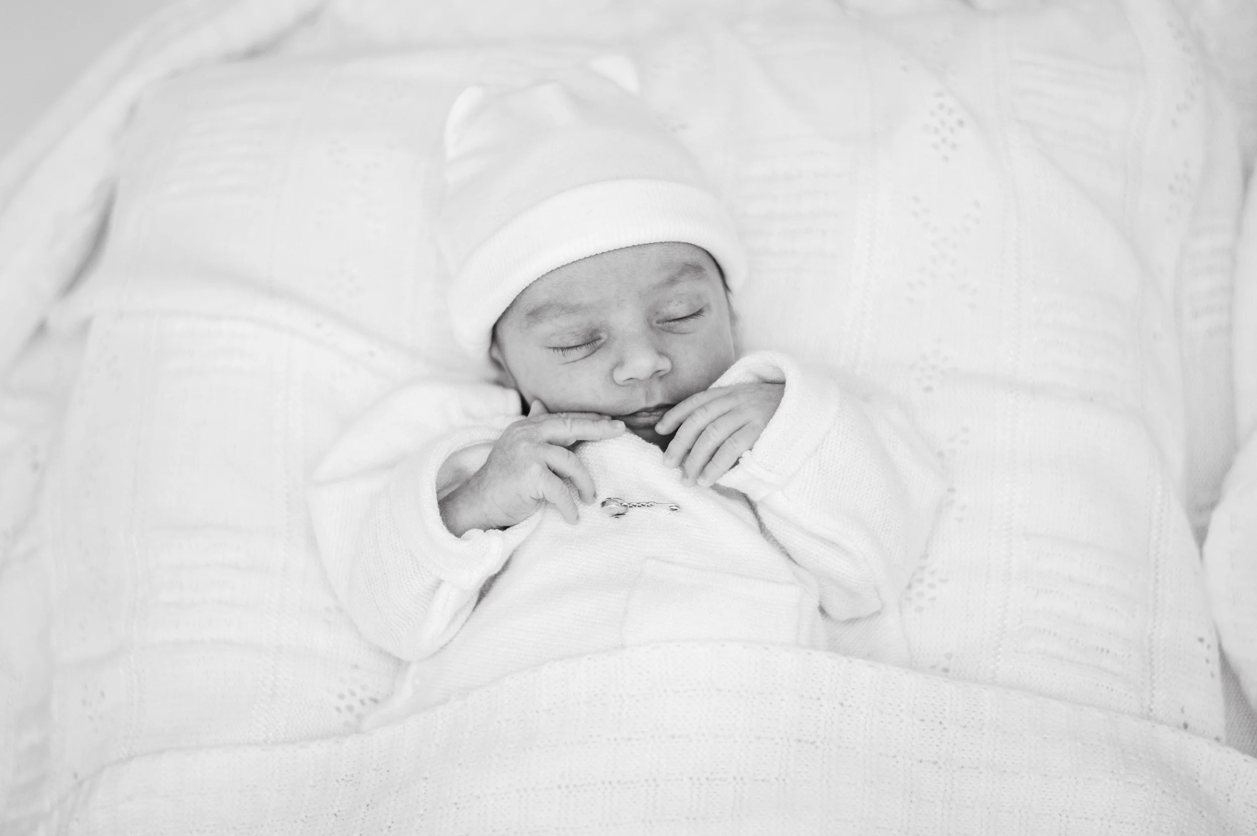Black and white photo of a newborn baby sleeping, wearing a hat and a long sleeve shirt, lying on a cushioned surface.