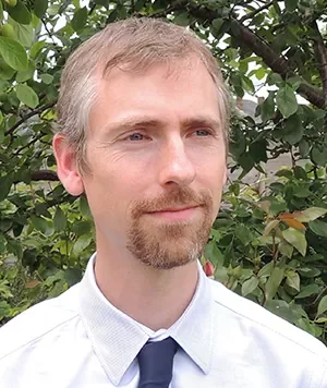 A man with light hair and facial hair wearing a white shirt and dark tie, standing outdoors in front of green foliage.