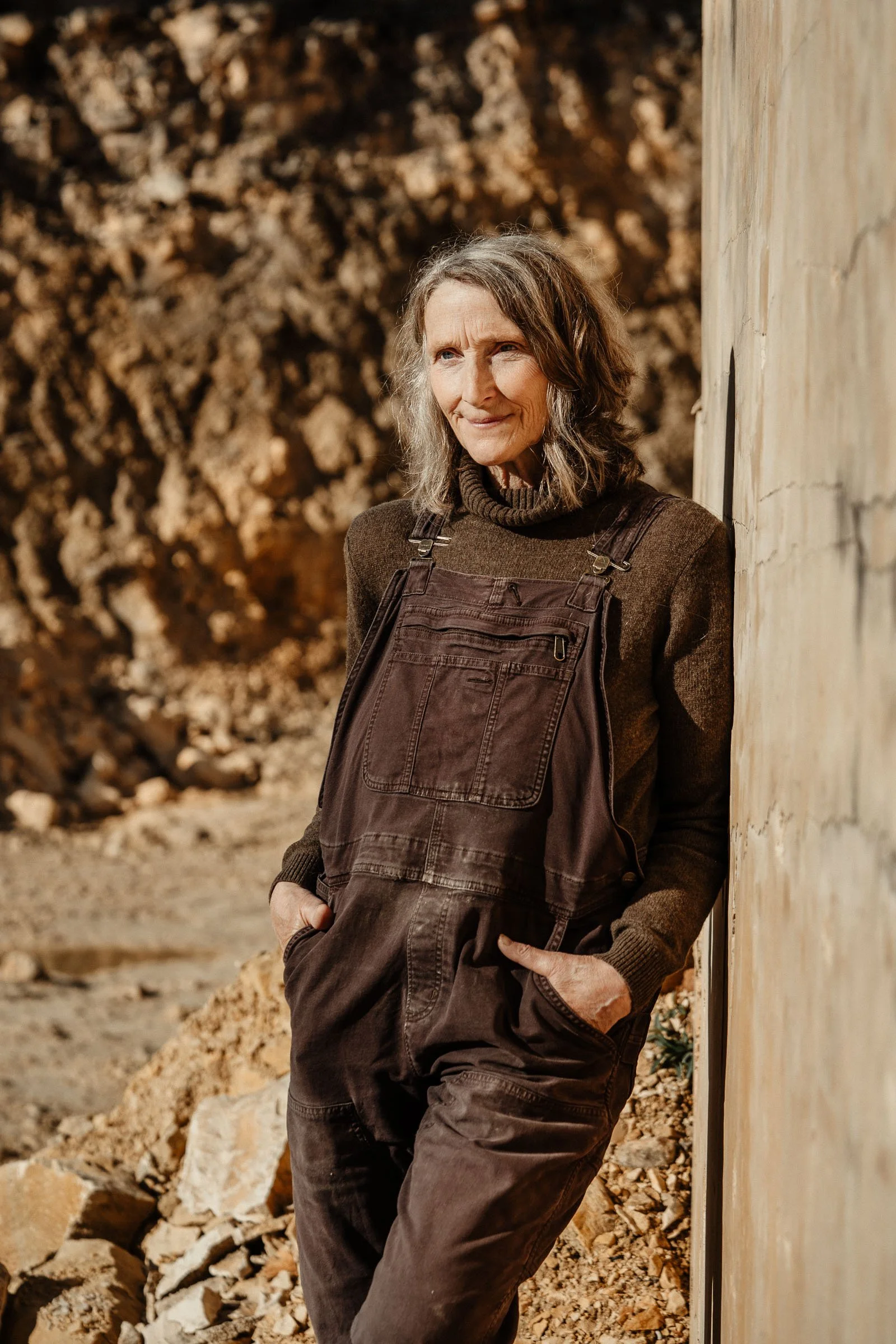 A woman with gray hair wearing brown overalls and a brown sweater, standing outdoors against a rocky background, leaning partially against a stone wall, with a slight smile on her face.