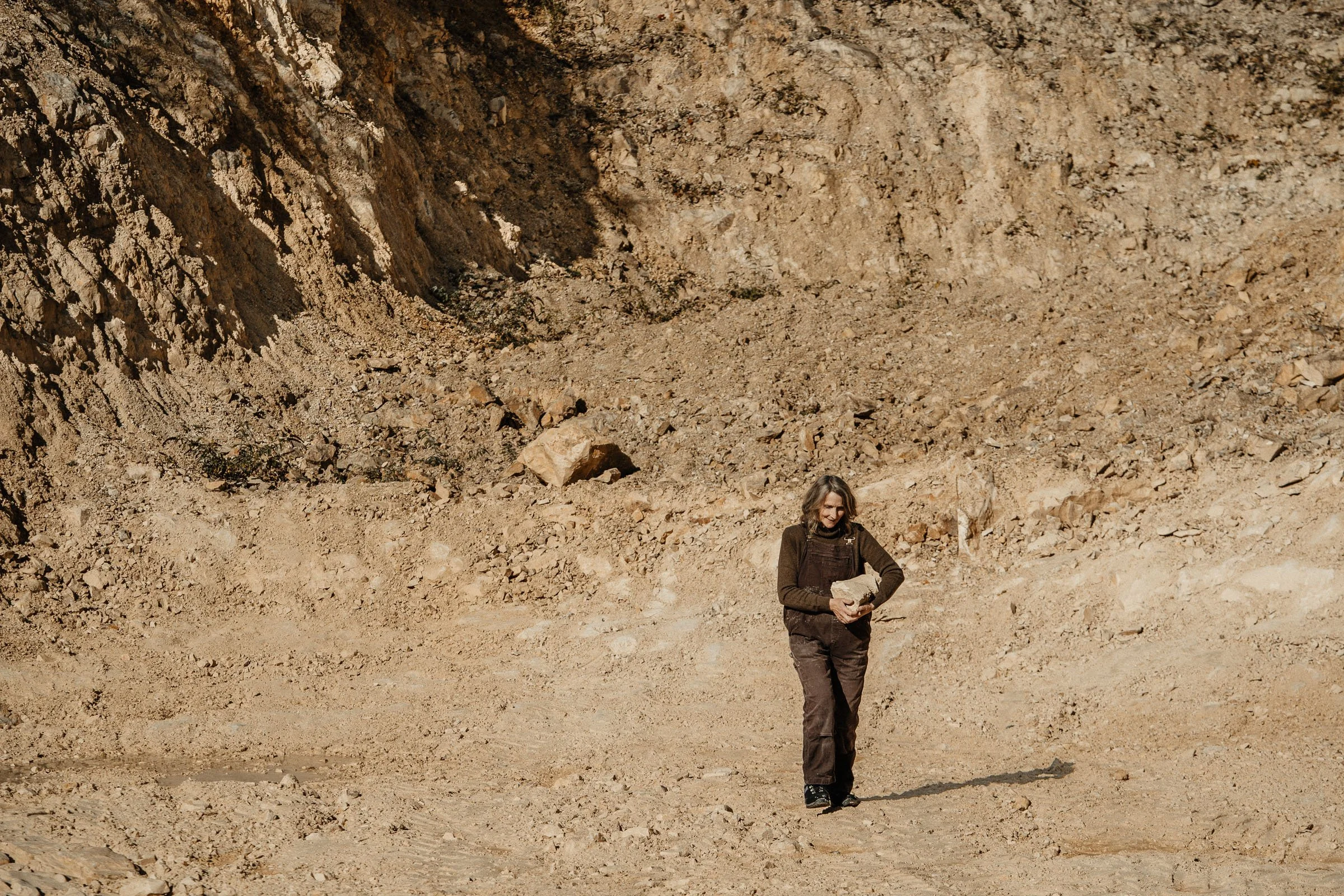 A woman walking in a rocky, desert landscape, holding a large rock in her hands, with a steep rocky cliff in the background.