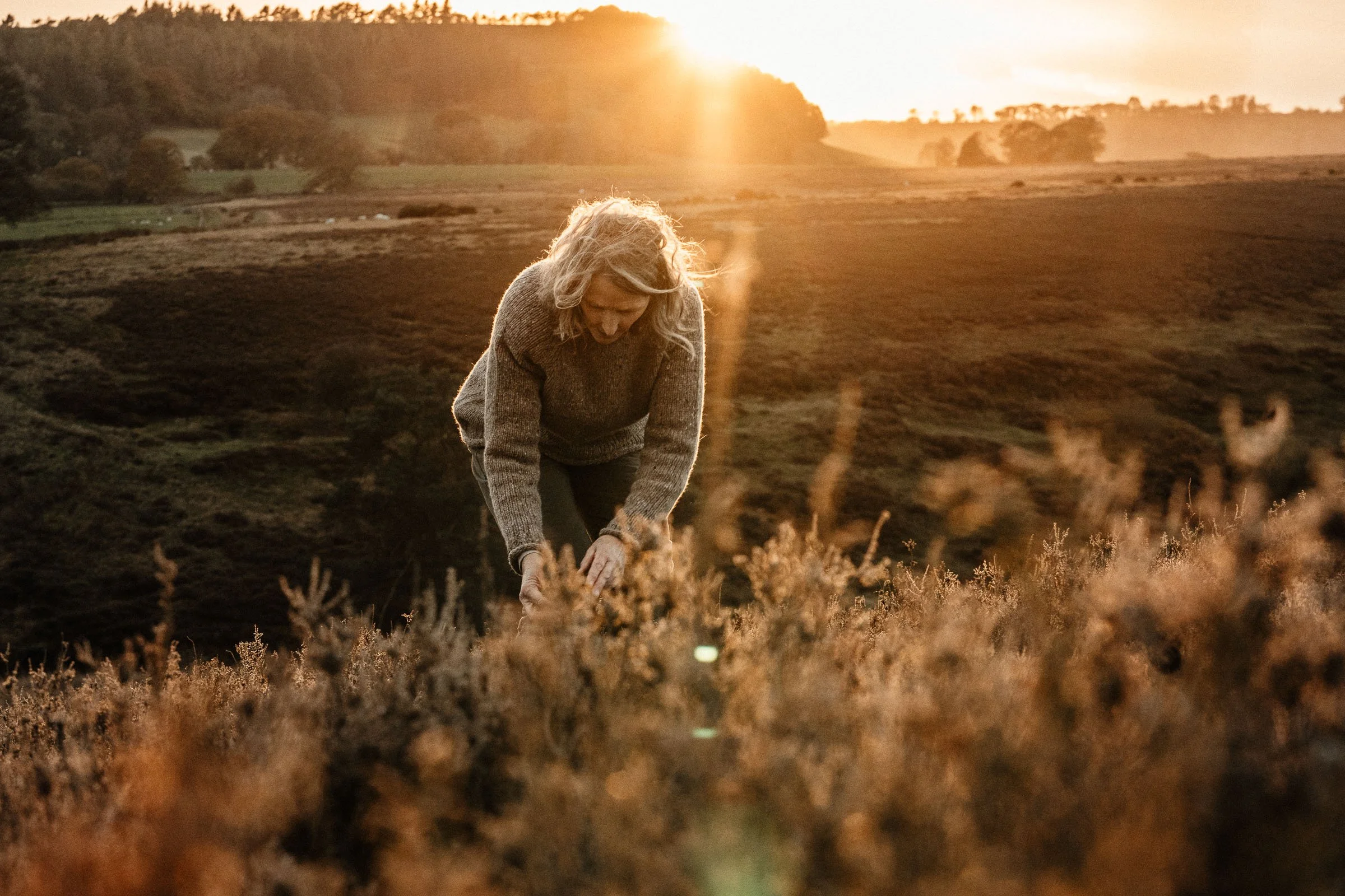 A woman in a cozy sweater bending down in a field during sunset, surrounded by tall grasses and rolling hills.