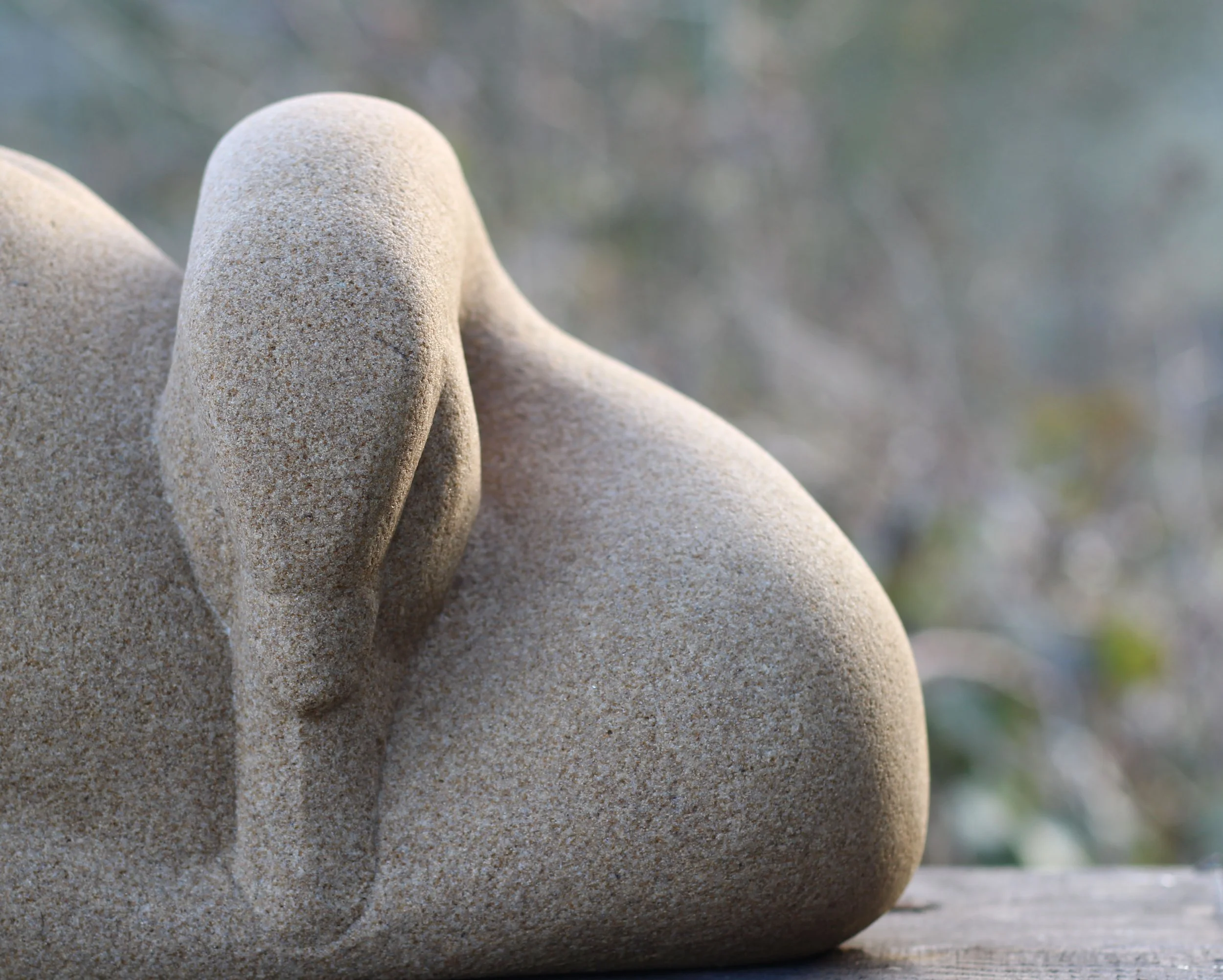 Close-up of a carved stone sculpture of a face with a hand covering one eye, on a wooden surface, with a blurred natural background.