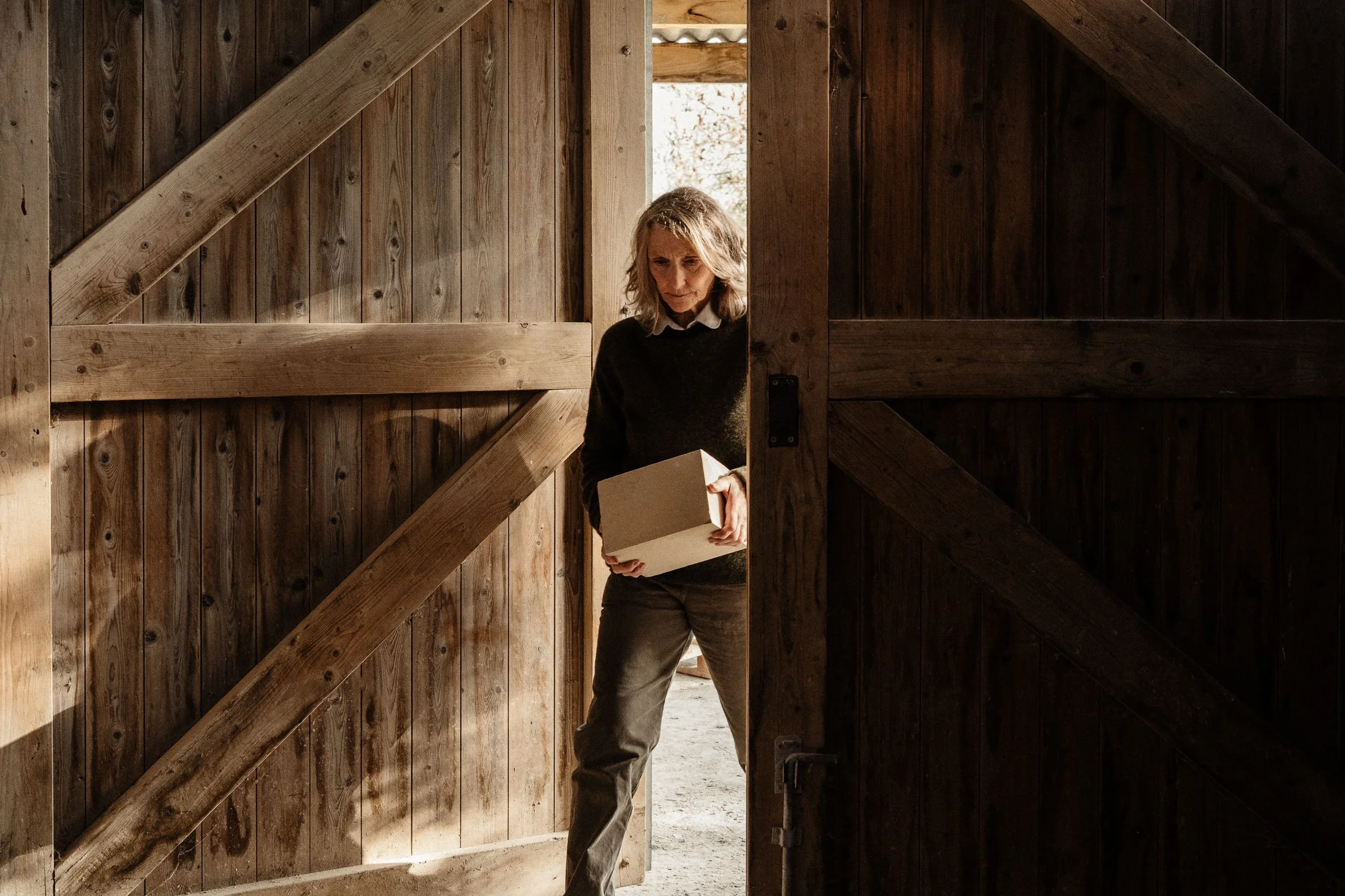 A woman standing in a wooden barn doorway, holding a cardboard box, with sunlit interior and wooden walls.