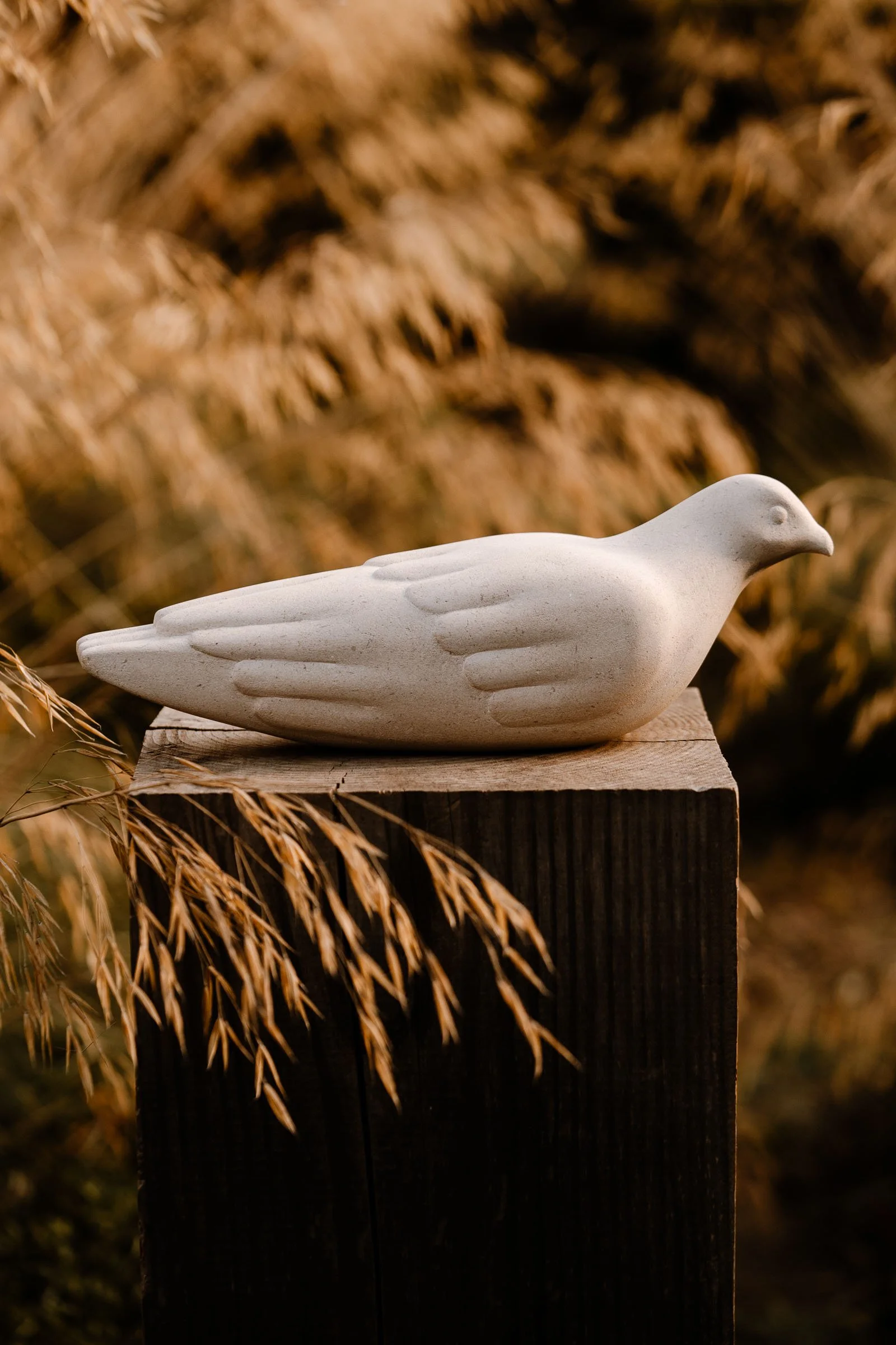 A white ceramic or stone dove sculpture placed on a wooden pedestal outdoors with dried brown plants in the background.
