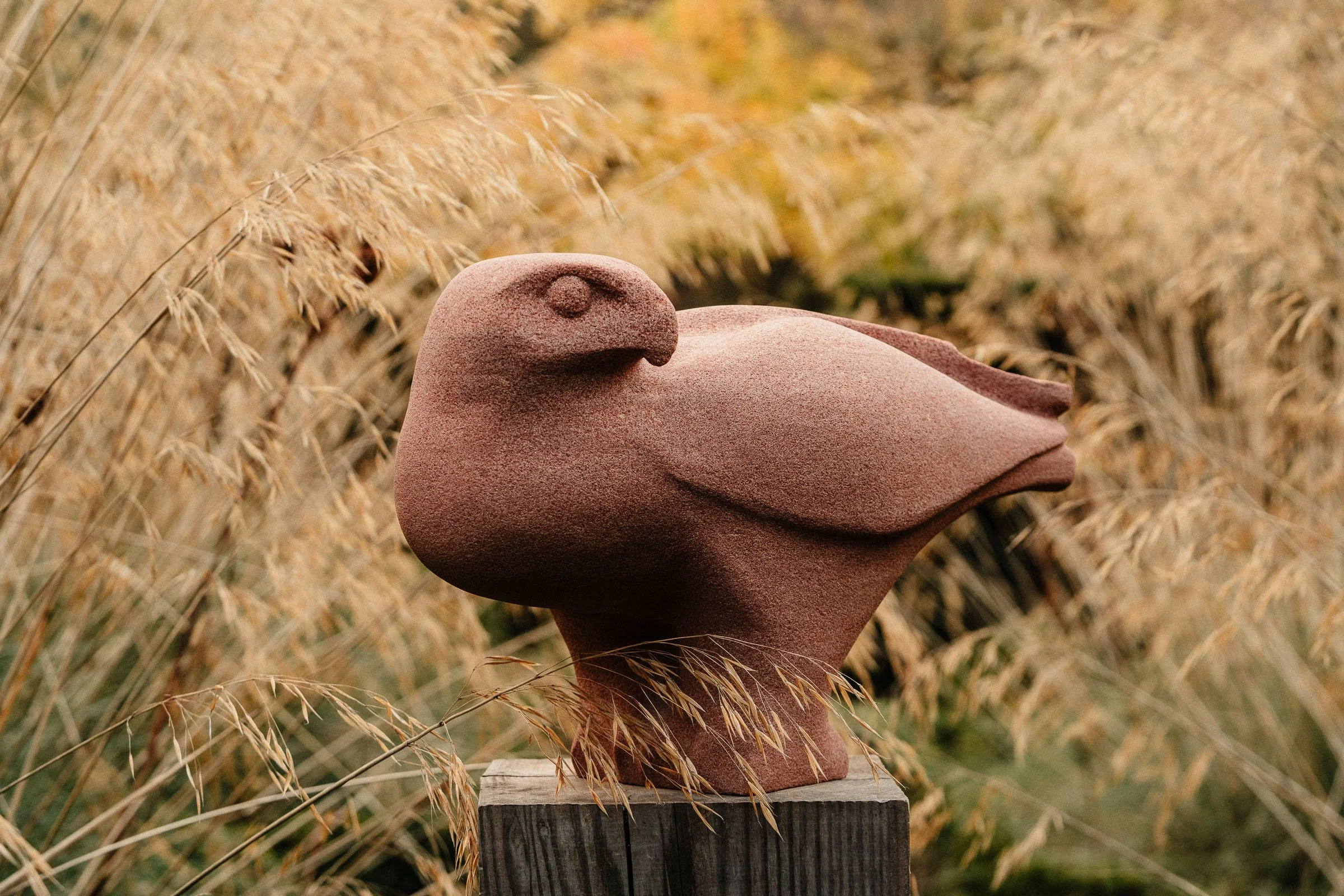 A stone sculpture of a bird, resembling a bird of prey, placed on a wooden pedestal amidst tall, dry grass.