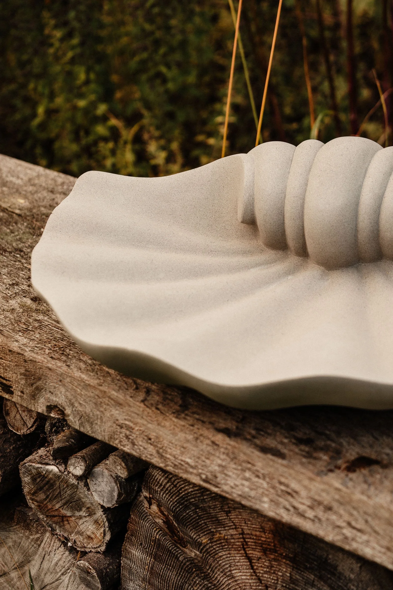 Close-up of a white ceramic dish with a wavy edge and ridges, placed on a rough wooden surface outdoors with a blurred green background.