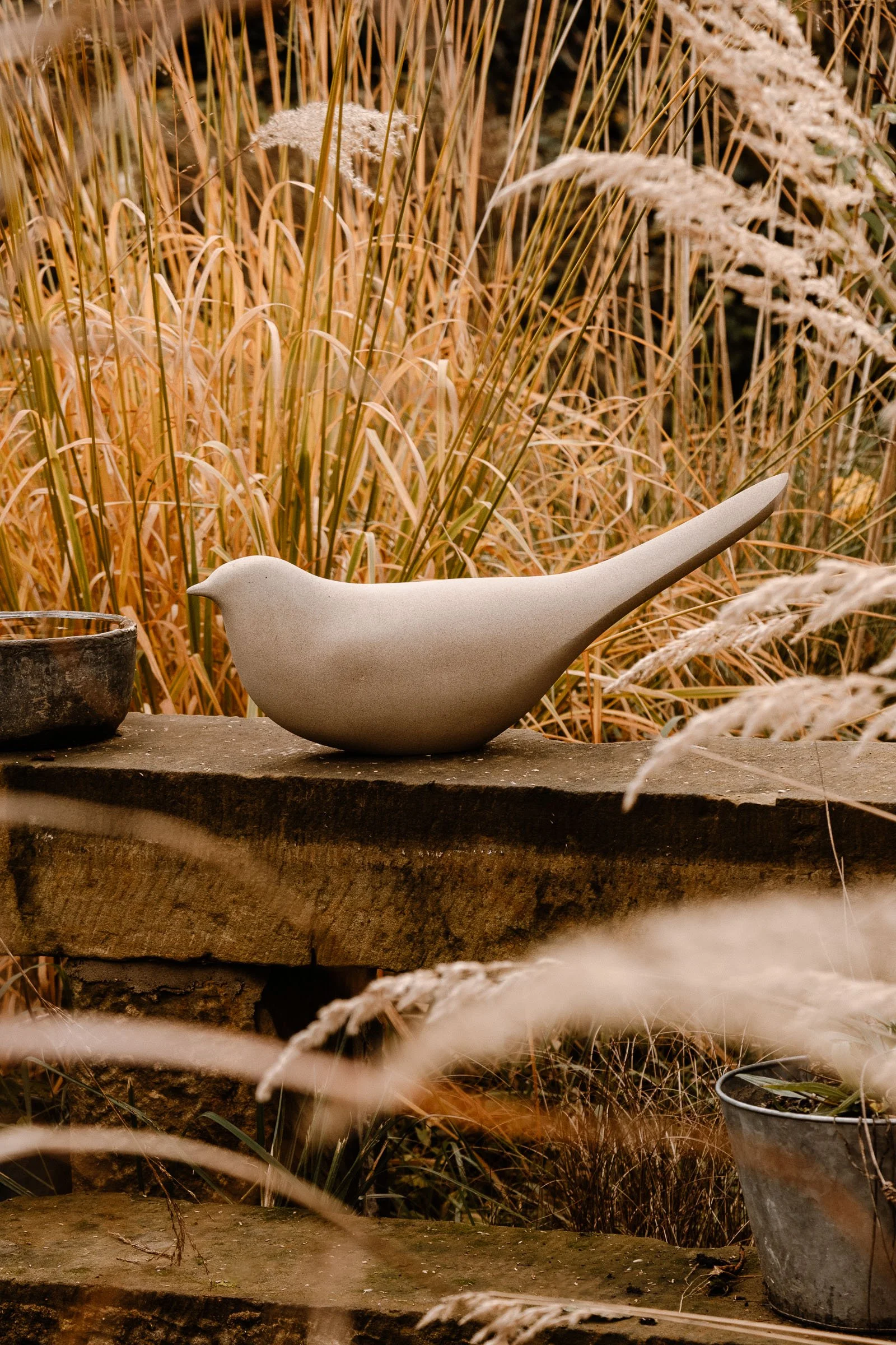 A decorative bird stone sculpture on a rustic wooden surface outdoors, surrounded by tall ornamental grasses and plants.