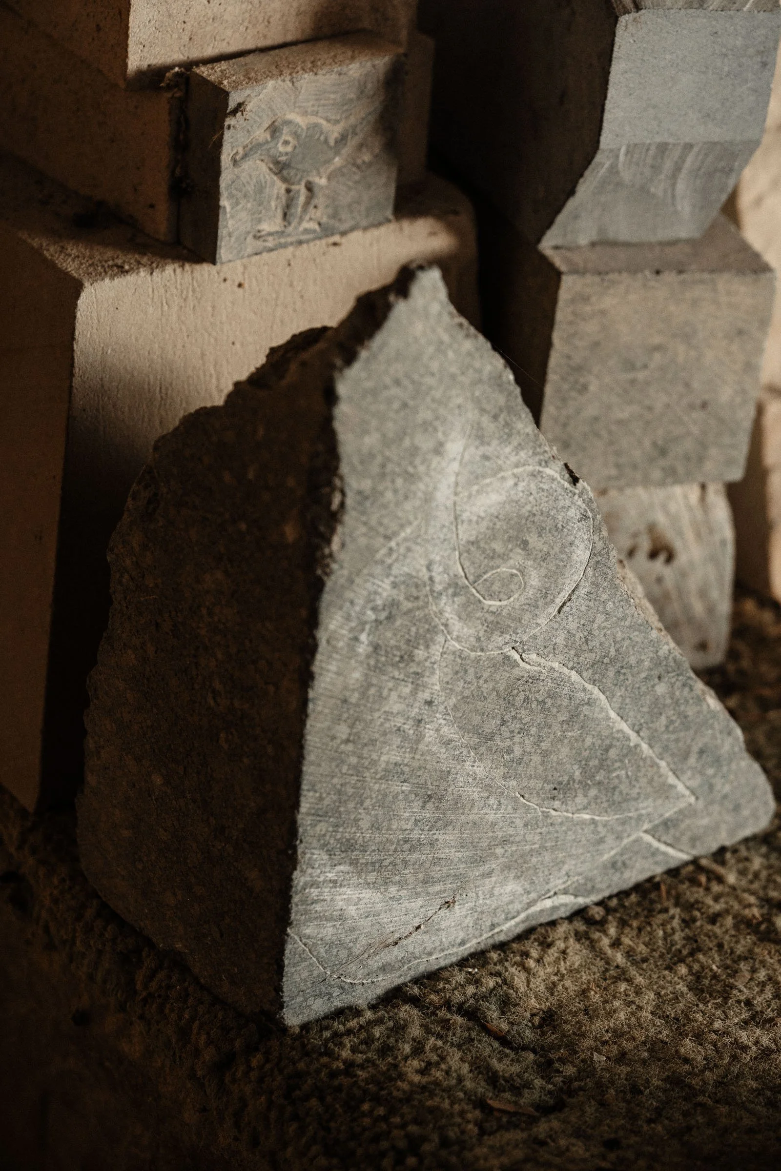 A close-up of a grey stone with a carved spiral and bird design, stacked among other stones in the sculpture studio.