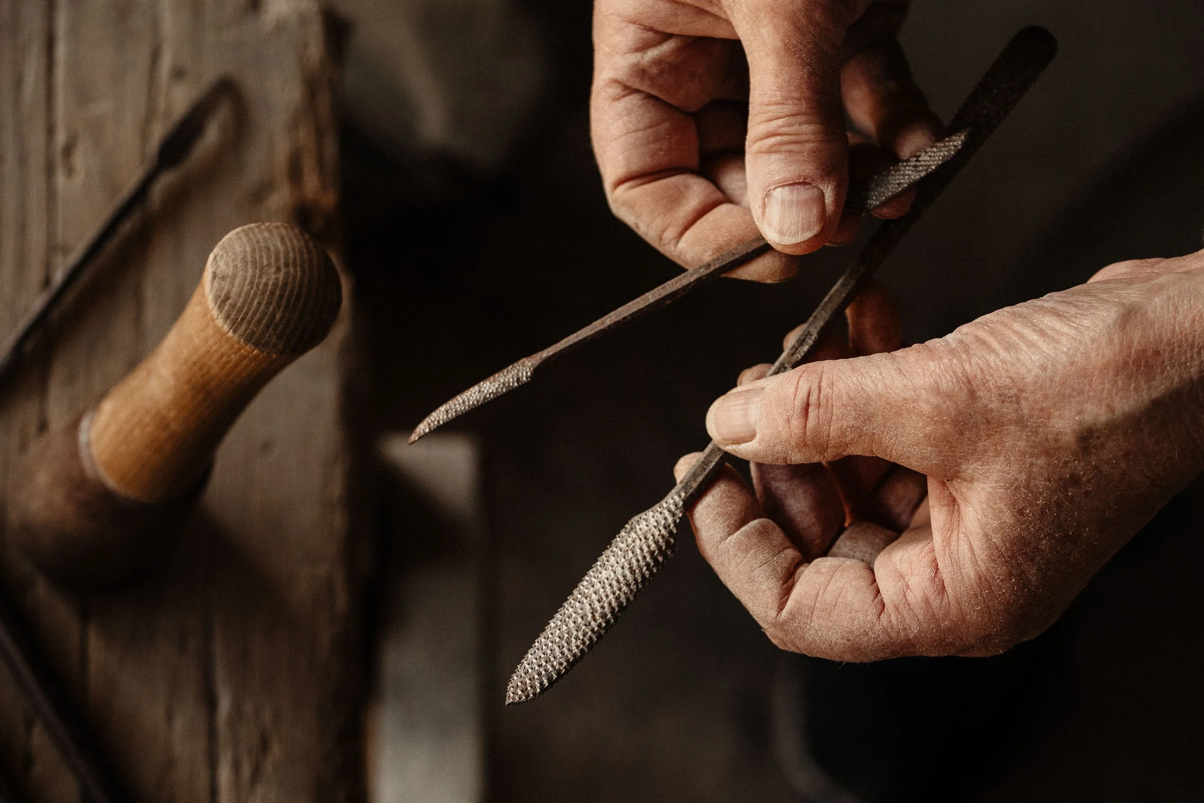 Close-up of a person's hands filing a small metal object with a metal file, with a wooden work surface and a wooden handled hammer in the background.
