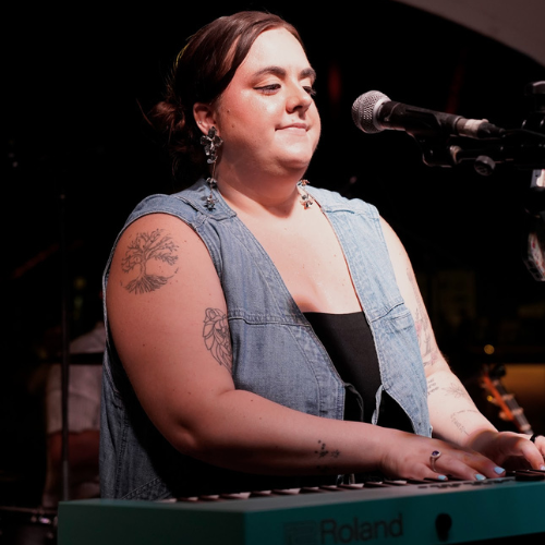 A woman with tattoos on her arms is playing a keyboard on stage, with a microphone nearby, in a performance setting.