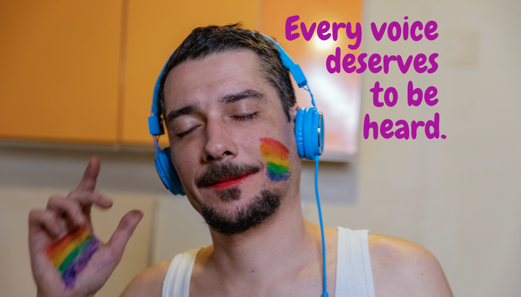 A man with rainbow-colored face paint and a rainbow-colored bracelet listening to music through blue headphones with eyes closed and a slight smile. The background is a room with yellow cabinets. The text on the image reads, "Every voice deserves to be heard" in purple font.