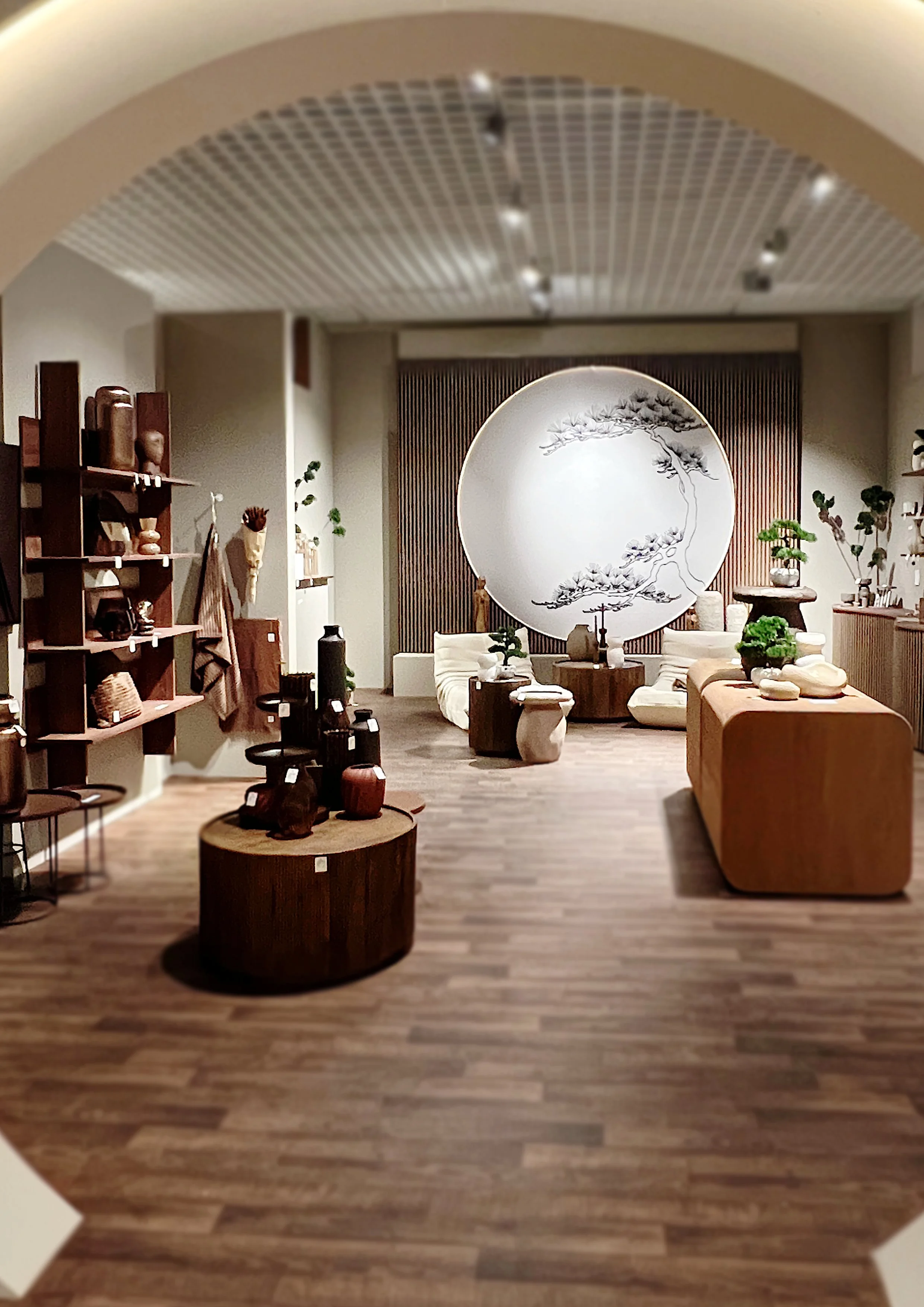 Ein stilvoll eingerichteter Raum mit asiatischem Dekor, einer großen runden Wanddekoration mit Baumzeichnung, Sitzecke mit Sofas, und Regale mit Pflanzen und Keramikartikeln.