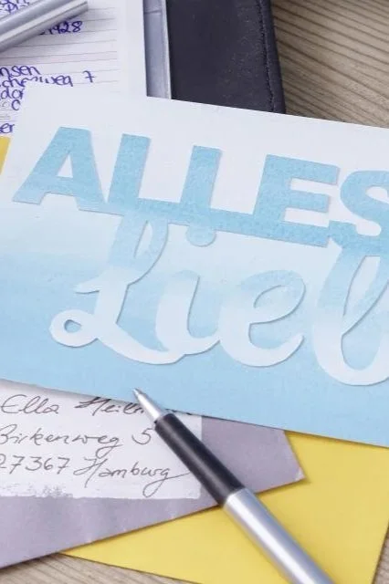 Weißer Briefbogen mit blauer Aufschrift 'ALLES Liebe', umgeben von Papier, Notizbuch und Kugelschreiber auf einem Tisch.