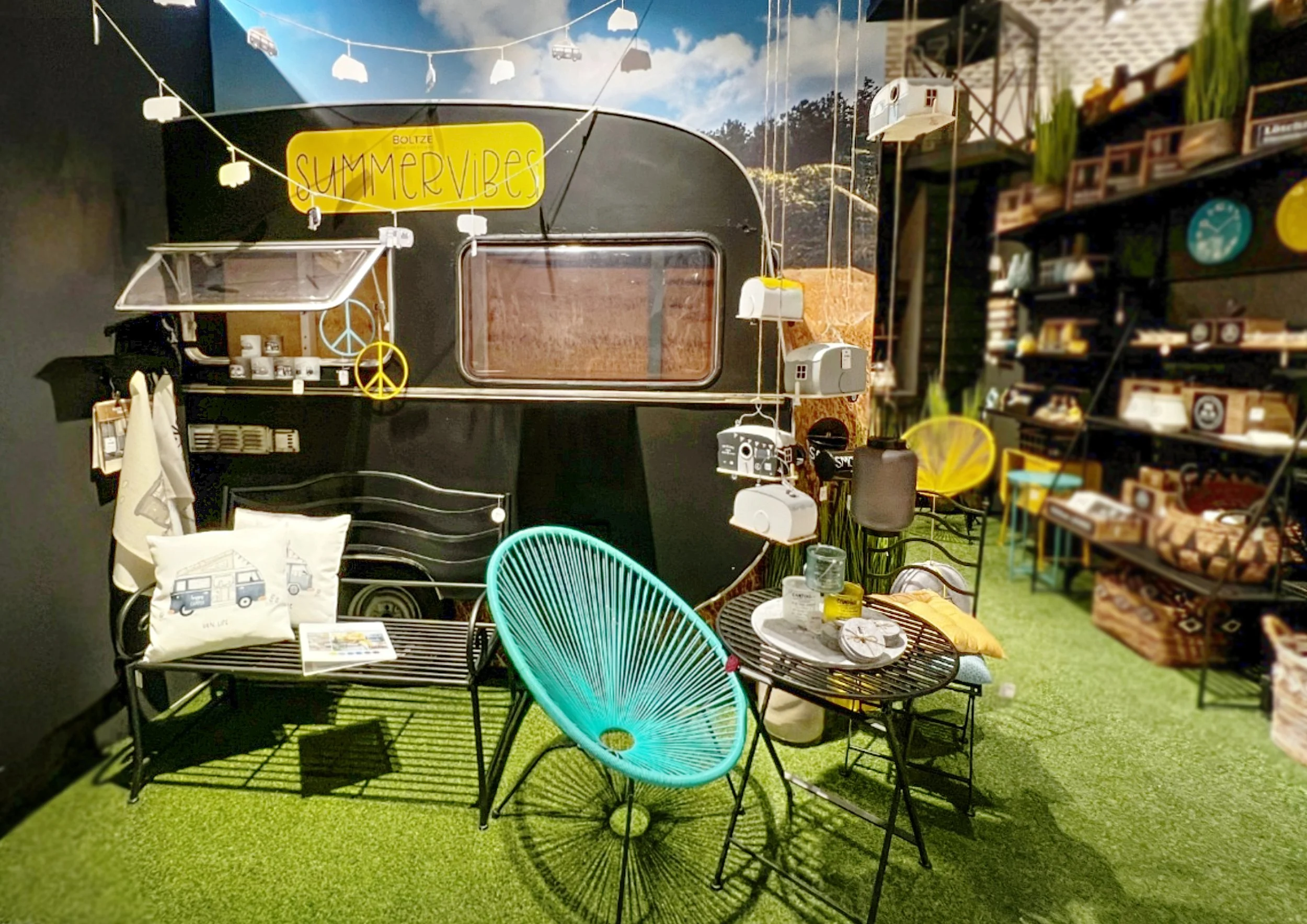 Ein Inneneinrichtung im Sommerstil mit einem schwarzen Wohnwagen, bunten Dekorationen, einem grünen Stuhl, einem kleinen Tisch mit Dekorationsartikeln und Regal mit Dekoartikeln auf einer grünen Teppichfläche.