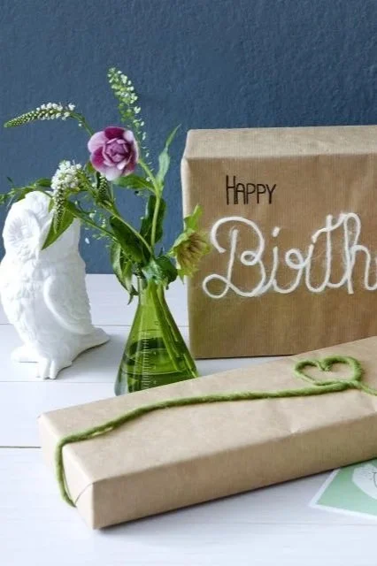 Ein Blumenstrauß in einer grünen Vase, eine Geschenkschachtel mit der Aufschrift 'Happy Birthday' und ein Geschenk in braunem Papier mit grünem Band, auf einem weißen Tisch gegen eine blaue Wand.