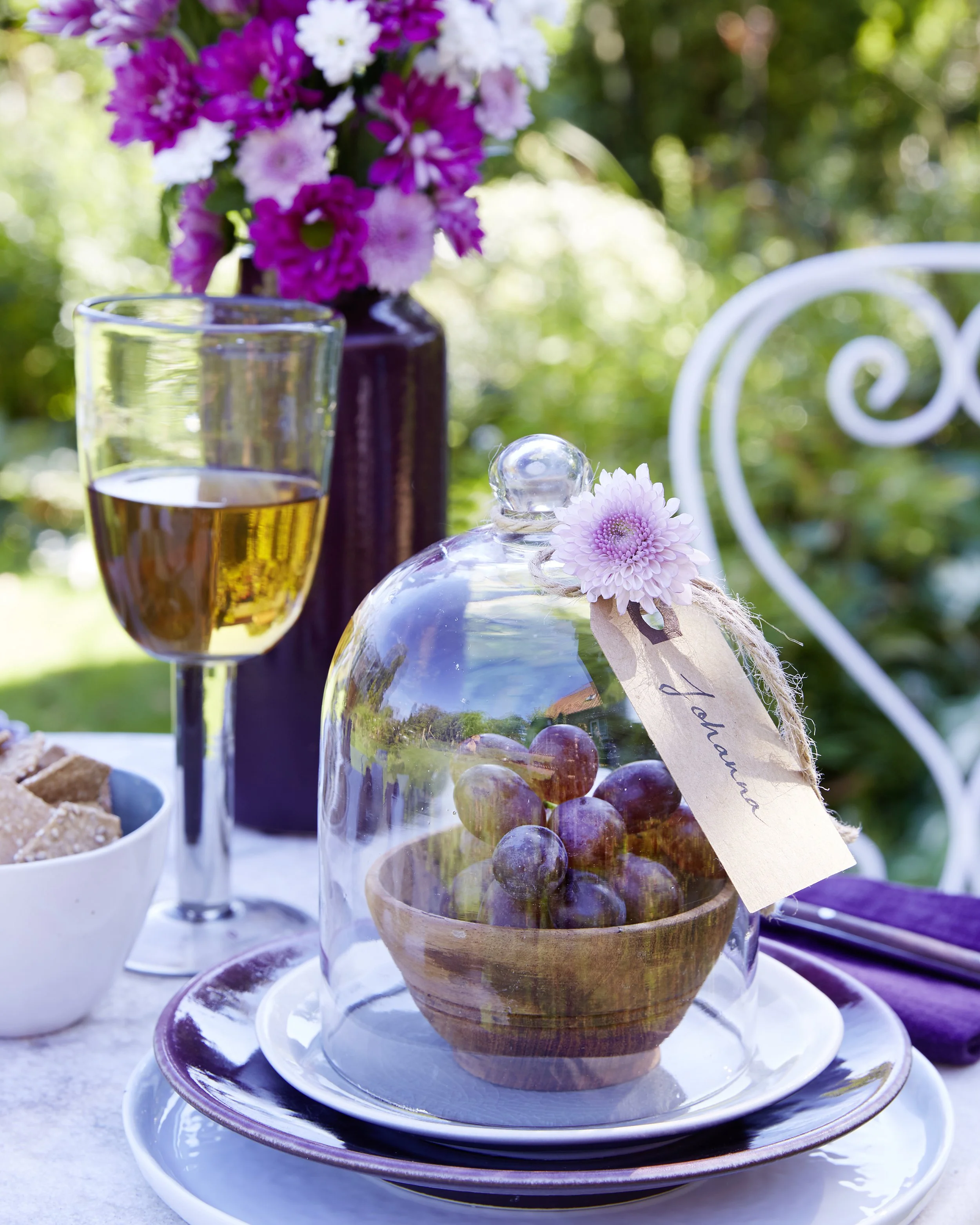 Ein stilvoll gedeckter Tisch im Garten mit einer Weintraube unter einer Glasglocke, einer Flasche mit pinken und weißen Blumen im Hintergrund, einem Glas Weißwein, einer Schale mit Keksen, einer weißen Gartenbank im Grünen und einem lila Tuch.