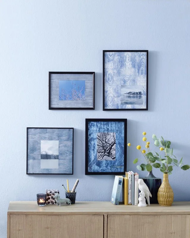 Ein Wandbild mit fünf Bilderrahmen in verschiedenen Größen und Motiven, darunter Wasserlandschaften, ein Baum und abstrakte Muster. Darunter ein Holztisch mit Deko-Objekten, Büchern und einer Vase mit grünen Zweigen mit gelben Blüten.