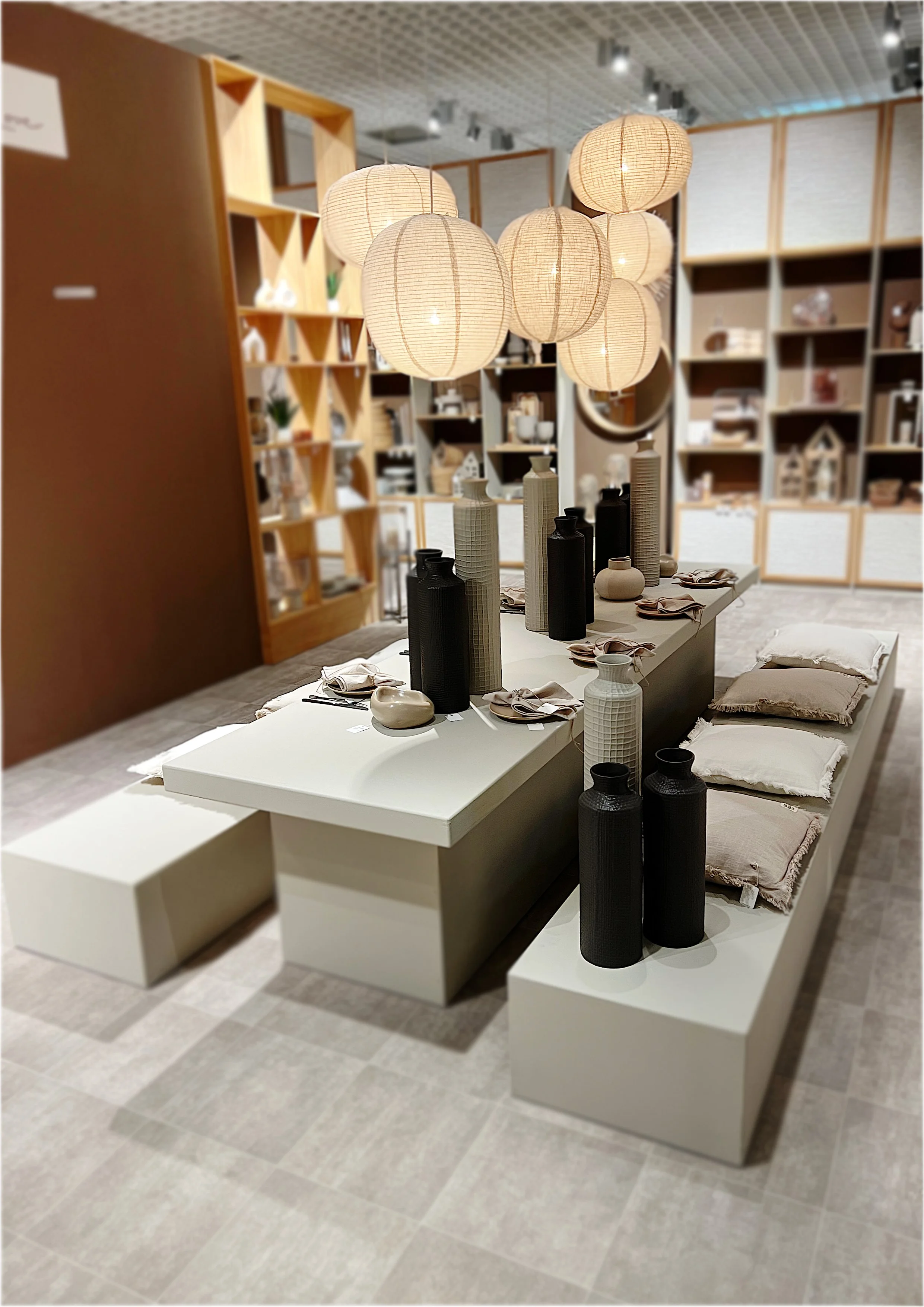 Deko-Tisch in einem Laden mit Lampions, Vasen und Kissen, umgeben von Regalen mit Geschirr und Dekorationsgegenständen.