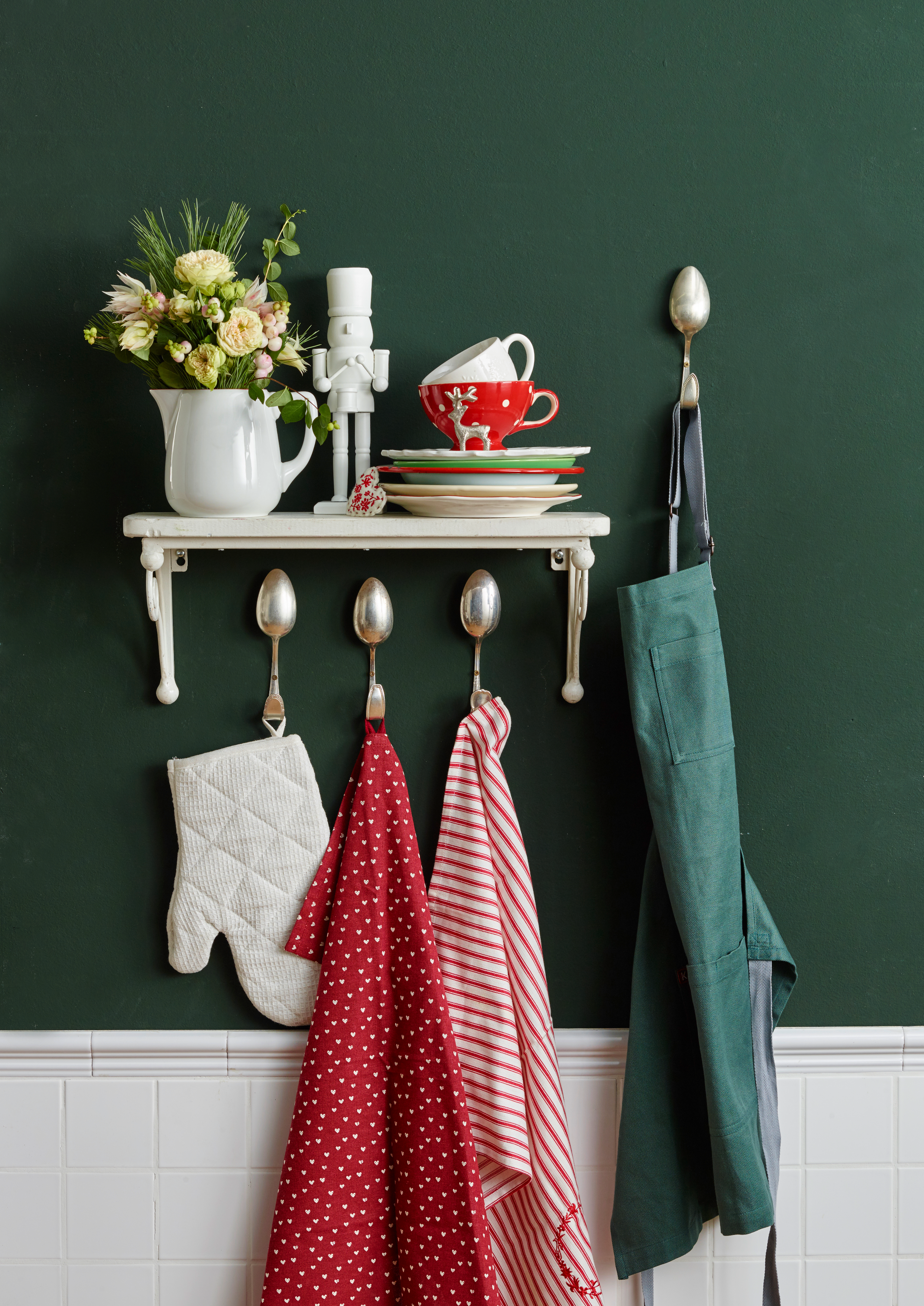 Weiße Küchenregal mit Blumen, Sektgläser, Teller, Tassen und Küchenutensilien vor einer grünen Wand.
