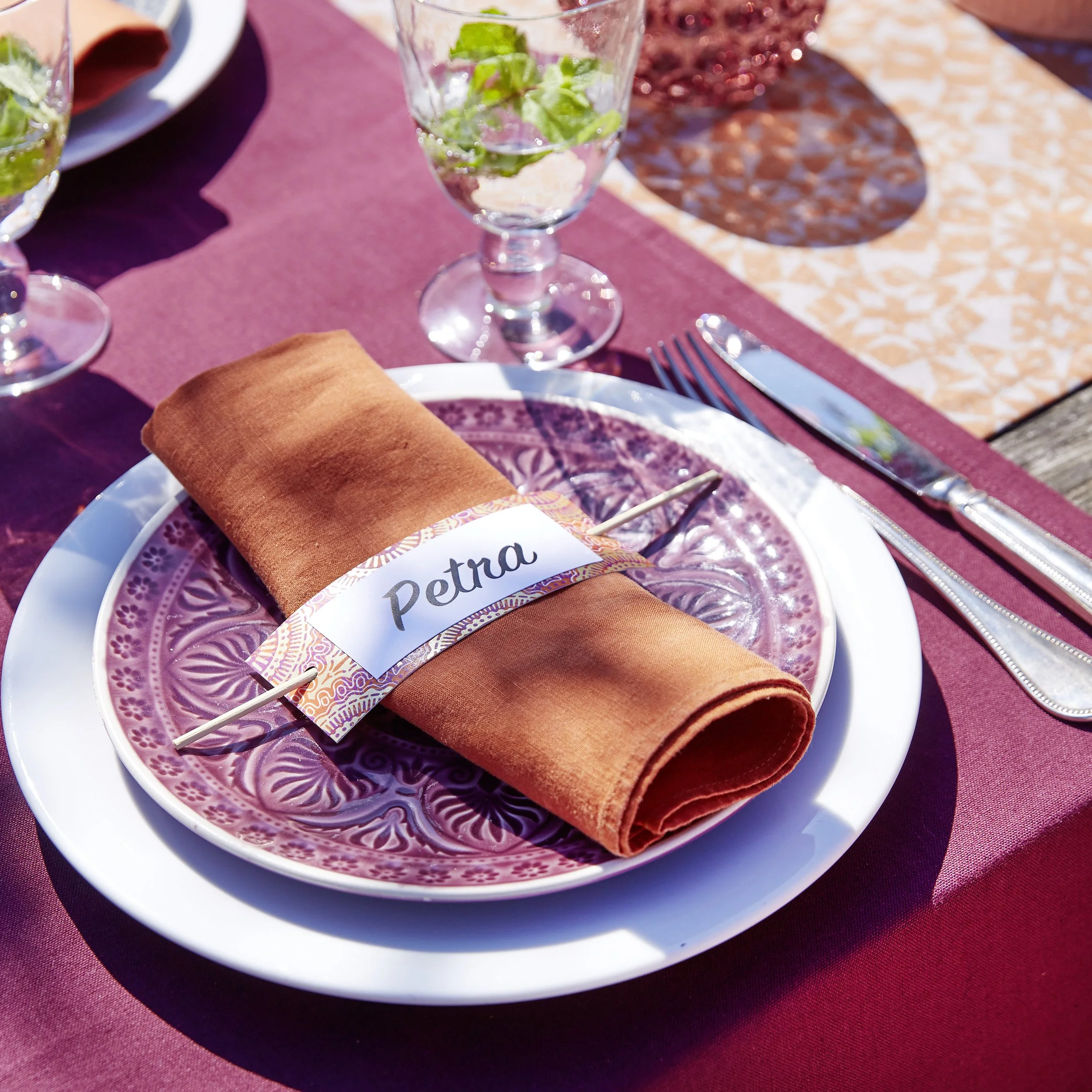 Ein gedeckter Tisch mit einem weißen Teller, einer lilafarbenen verzierenden Untertasse, einem goldenbraunen Stoffserviette, einem Namensschild mit 'Petra', Trinkgläser mit Wasser und Minzblättern, und Besteck auf einem roten Tischläufer.