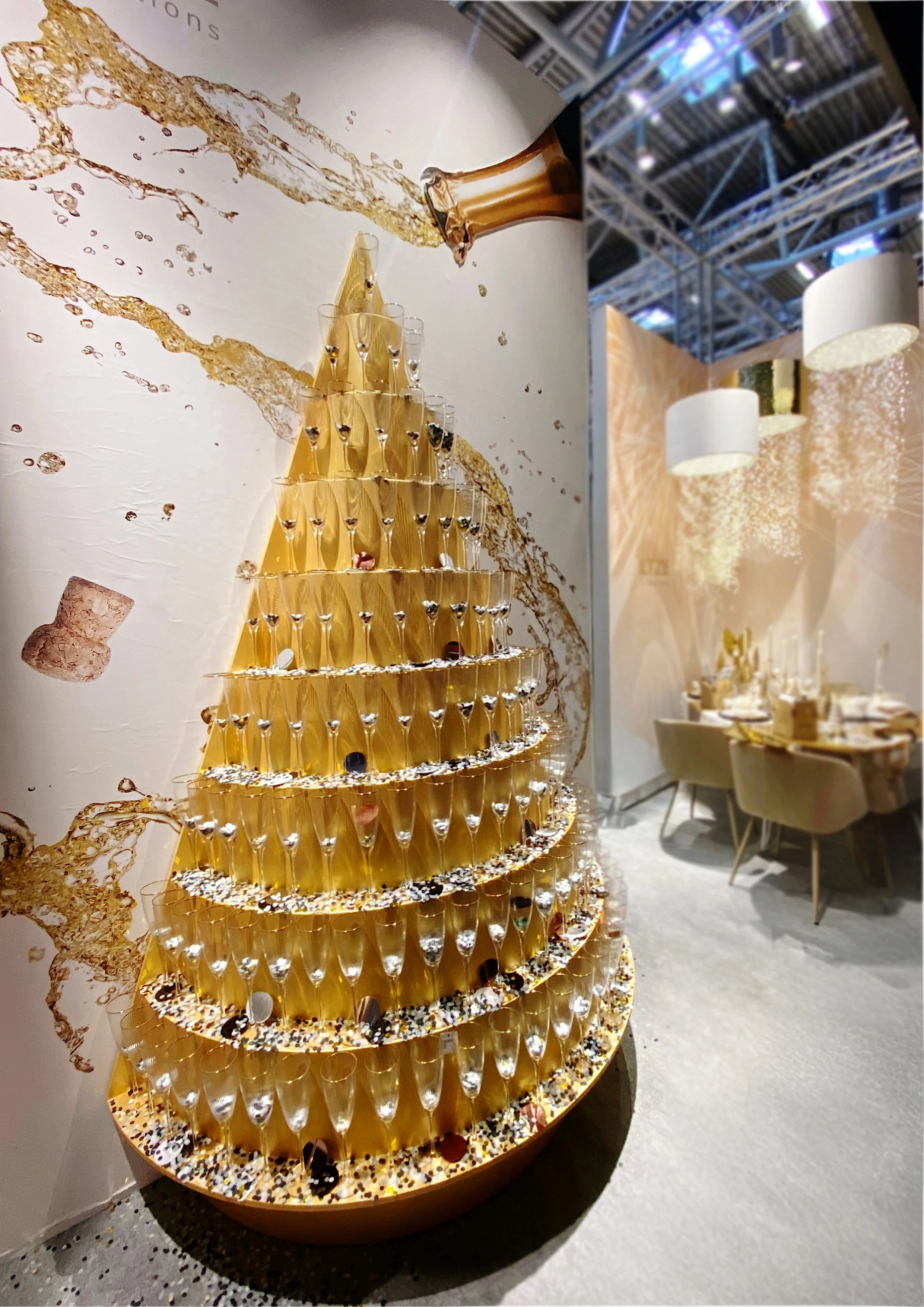 Ein dekoratives Kunstwerk in Form eines Goldbaums bestehend aus mehreren Schichten voller Gläser, die wie Champagner aussehen, mit Konfetti und Dekorationen am Boden, vor einer Wand mit Champagner- und Bubble-Design. Im Hintergrund ein festlich gedeckter Tisch mit Stühlen.
