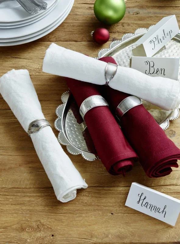 Weihnachtlicher Tisch mit verbundenen Stoffservietten in Weiß und Rot, dekorative Serviettenring, Name-Schilder, und festliche Dekorationen wie kleine farbige Kugeln.