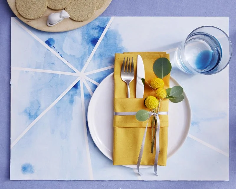 Gedeckter Esstisch mit gelbem Stoffservietten, Besteck, kleinen gelben Blumen, klarem Wasser, und einem blauen Wasserglas auf Esstisch.