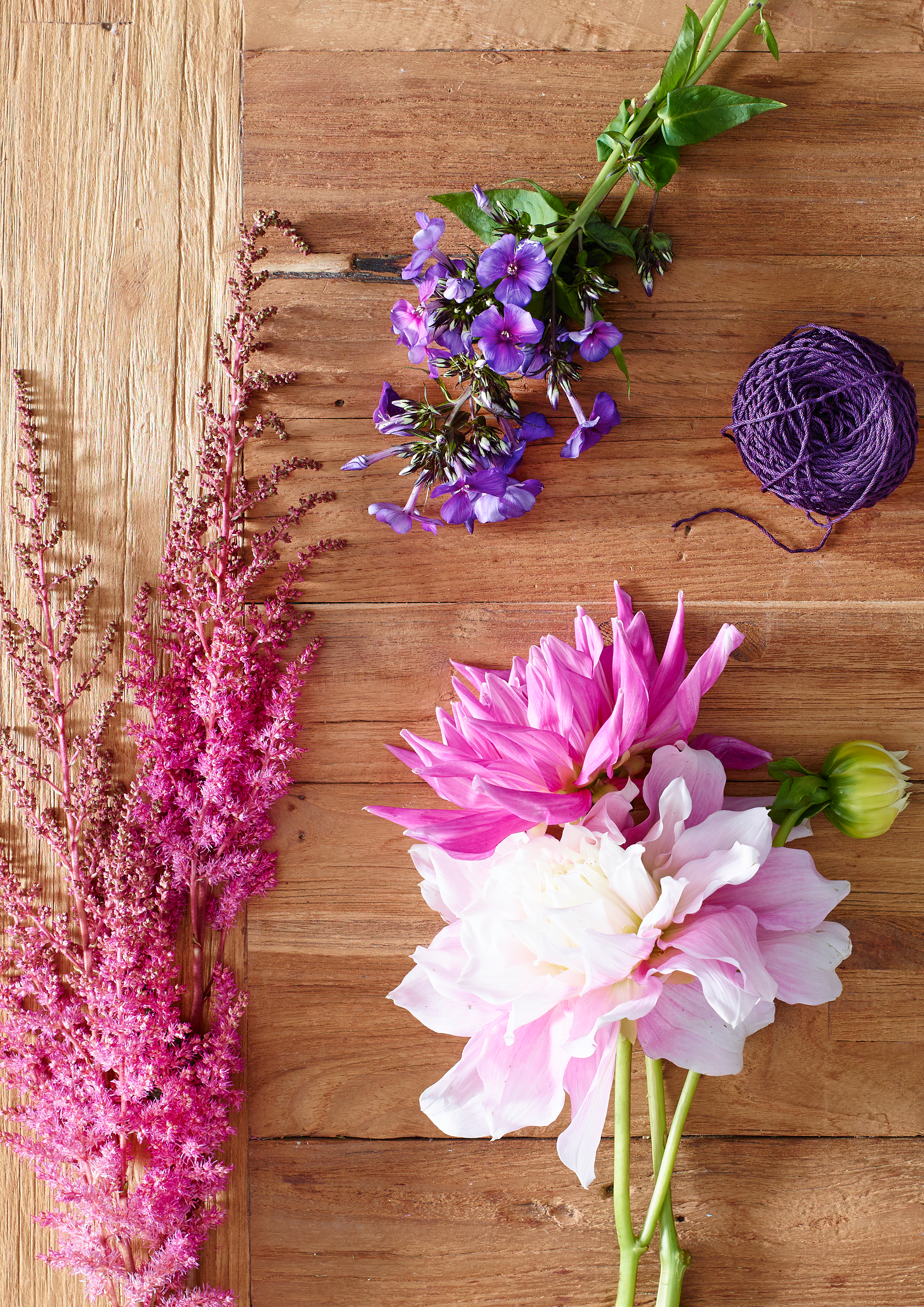 Verschiedene Blumen, darunter pink-weiße Dahlien, lila Glücksklee, pinke Astilbe und ein grünes Knospen. Ein schwarzes Wollknäuel auf einem Holztisch.