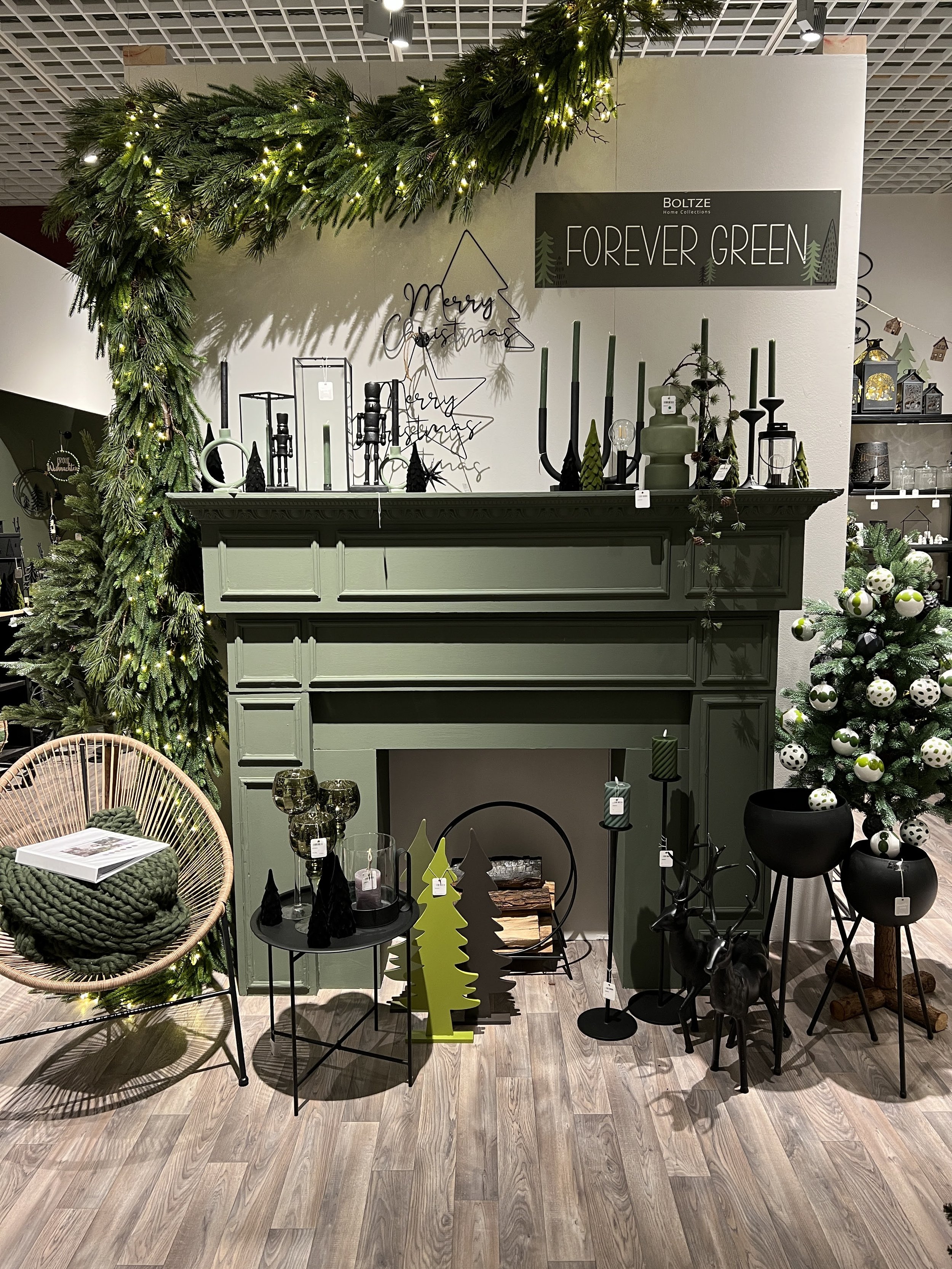 Weihnachtliche Dekorationen in einem Möbelgeschäft, darunter ein grüner Kamin, Weihnachtskugeln, Kerzen, künstliche Tannenbäume und verschiedene stilvolle Dekorationsartikel.