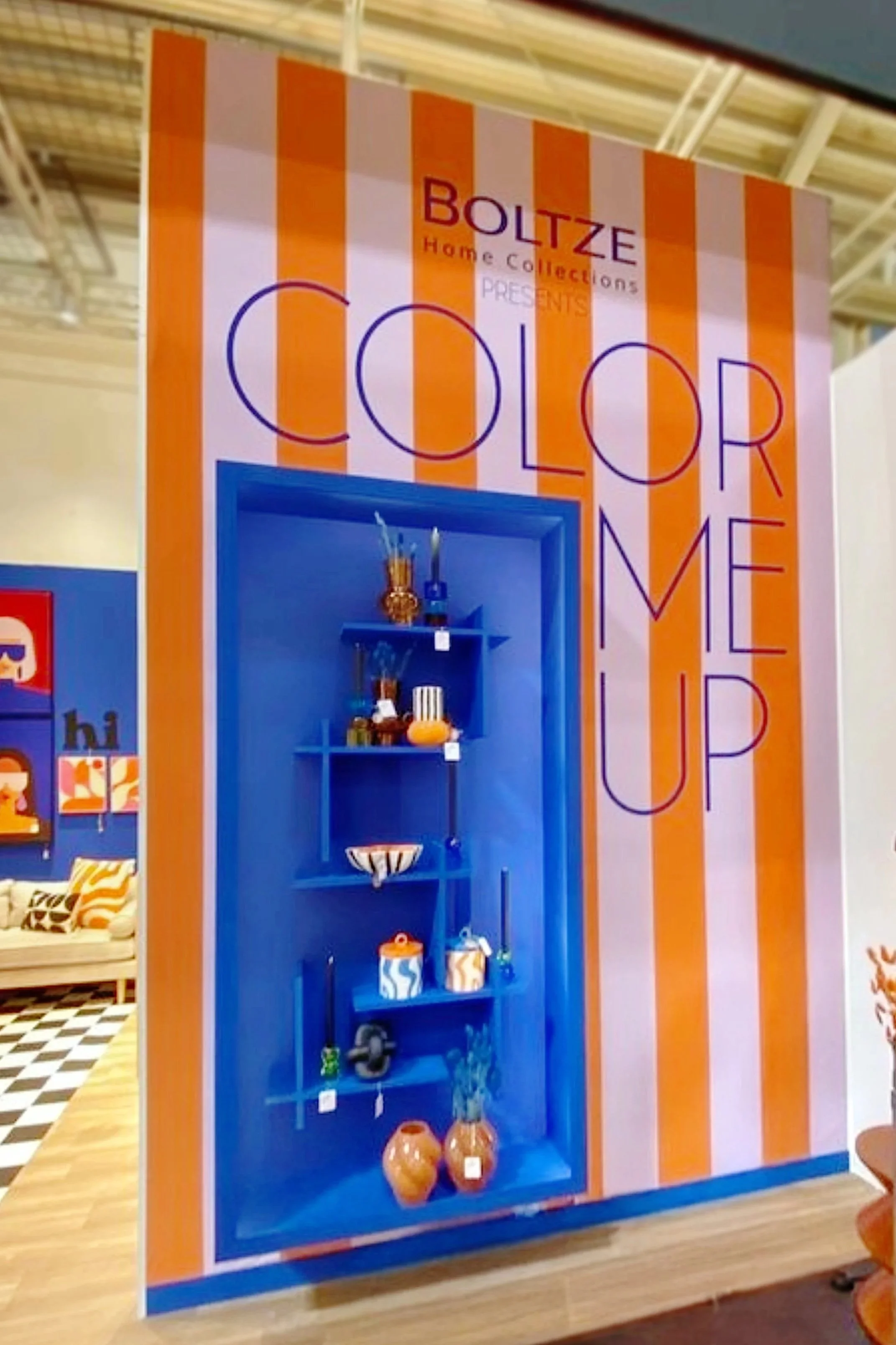 Ein Werbeplakat mit orange-weißen Streifen und dem Text 'BOLZ Home Collections präsentiert COLOR ME UP' sowie eine blaue Regalwand mit verschiedenen Dekorationsgegenständen.