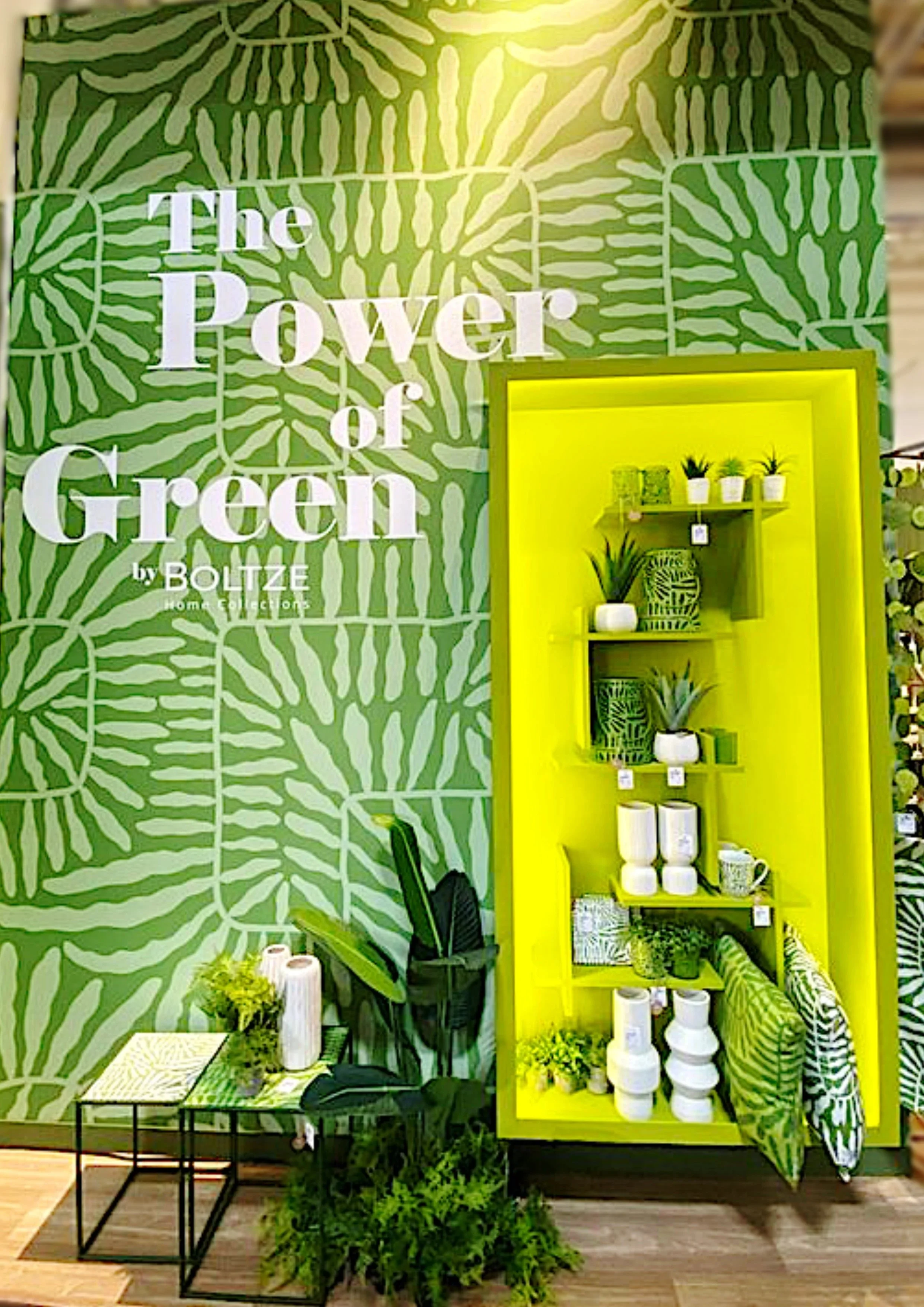 Eine grüne Wand mit Palmblatt-Design, daneben ein gelb gestrichener Regal mit Pflanzen und Dekorartikeln. Im Vordergrund befinden sich zwei kleine Tische mit Pflanzen und Vasen.